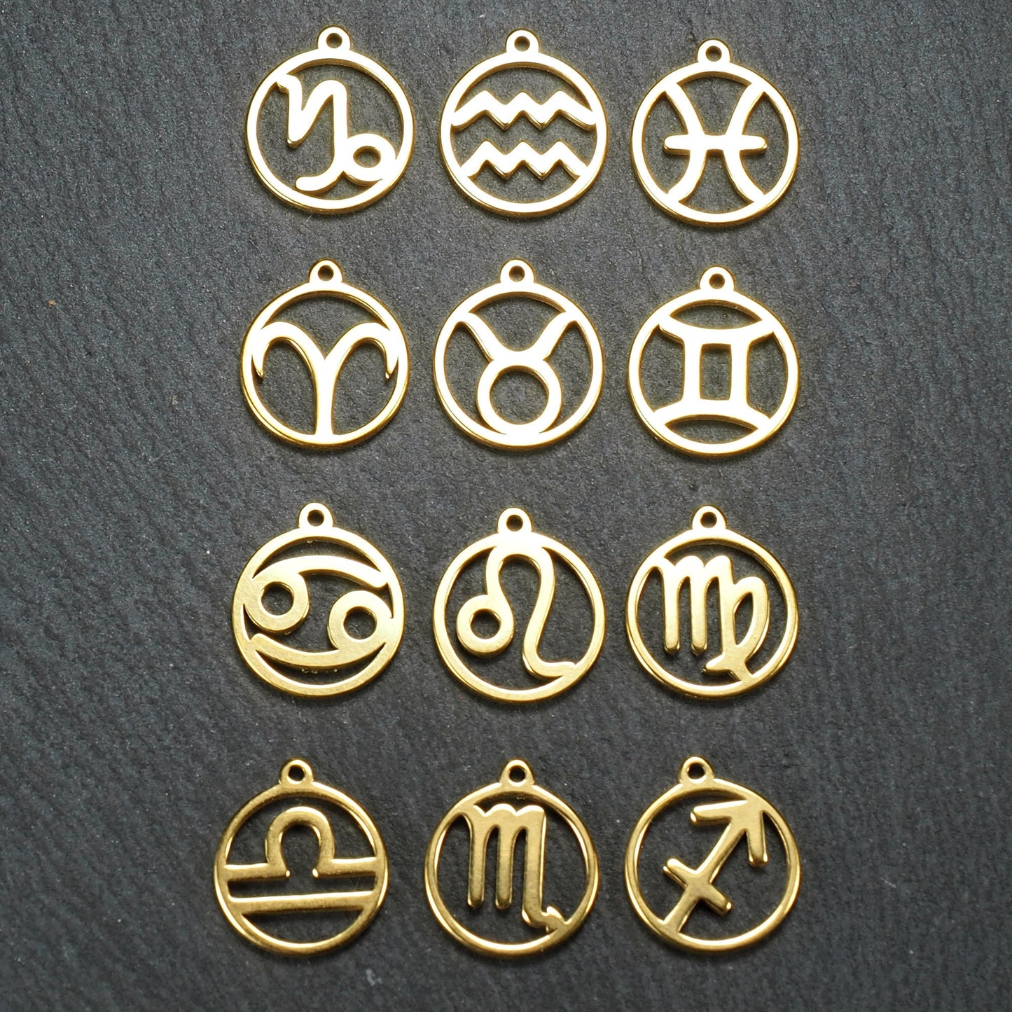 Zwölf goldfarbene, glänzende, runde Anhänger mit durchbrochenen Sternzeichen-Symbolen in feinem Metallrahmen, jeweils mit kleiner Öse oben.