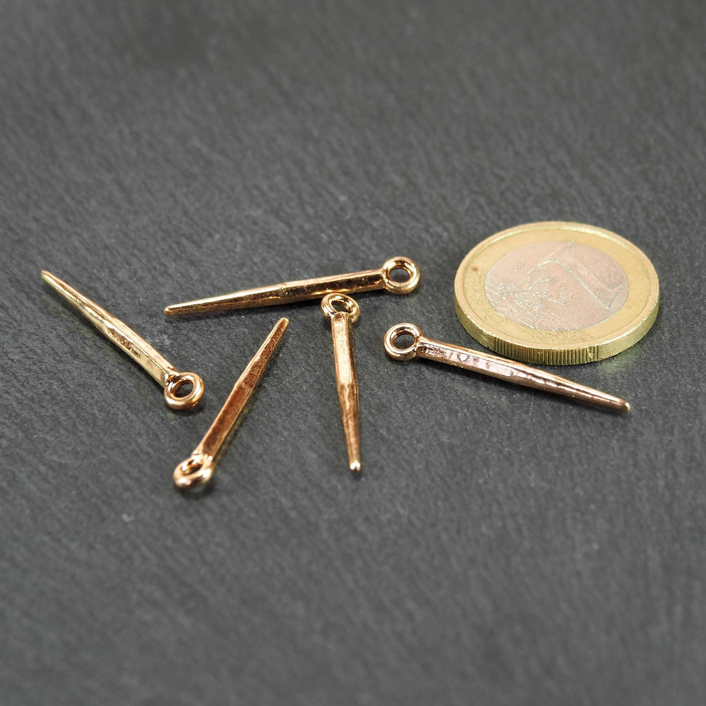 Fünf goldfarbene, konische Anhänger mit sichtbarer Öse und glänzender Oberfläche, spitz zulaufend, neben einer 1-Euro-Münze.