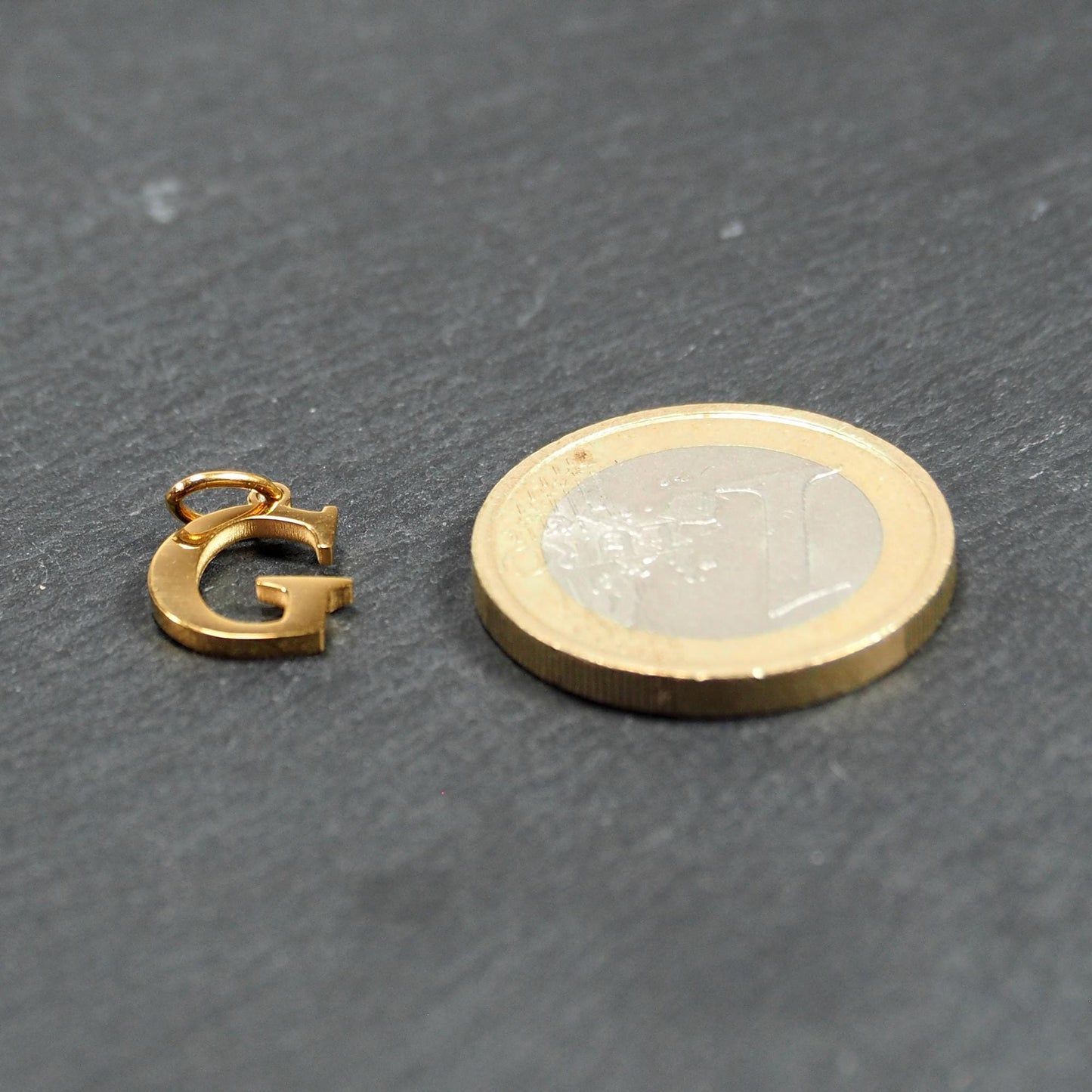 Goldfarbener, glänzender Buchstabenanhänger in Versal-Serifenschrift (Großbuchstabe G) mit kleiner Öse, neben einer 1-Euro-Münze zur Größendarstellung.