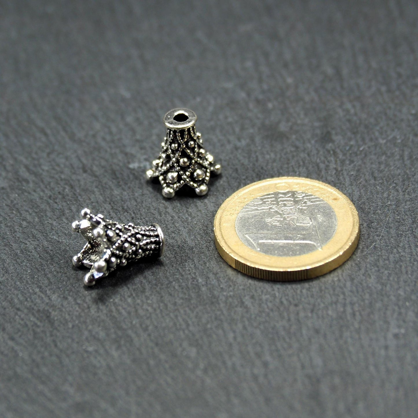 Zwei konische, antik-silberne, durchbrochene Perlenkappen mit filigranem Relief und kleinen Noppen, neben einer 1-Euro-Münze zur Größenanzeige.