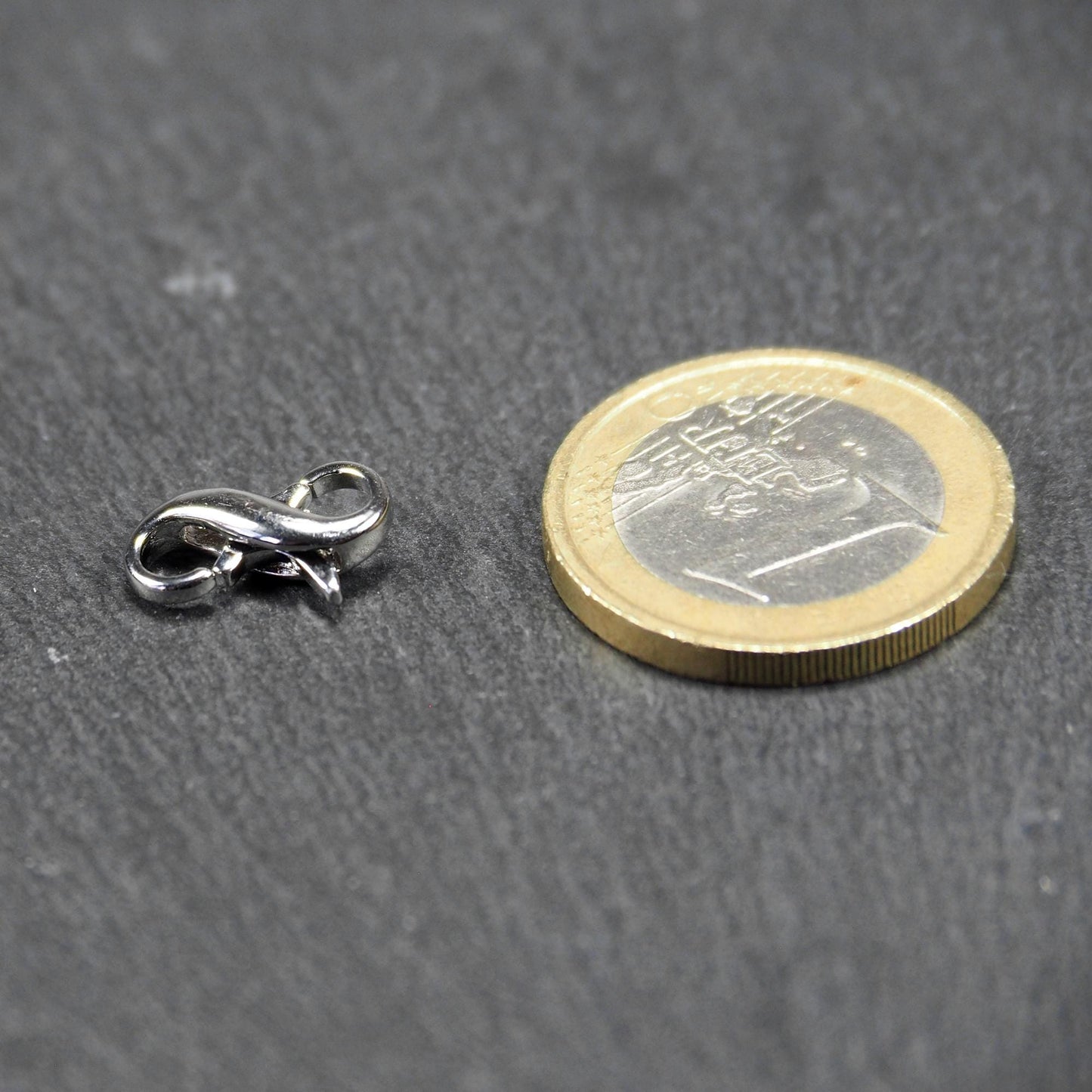 Ein doppelseitiger Karabiner im liegenden Acht-Design, silberfarben glänzend mit organisch geschwungener, durchbrochener Form und integrierter Öse.