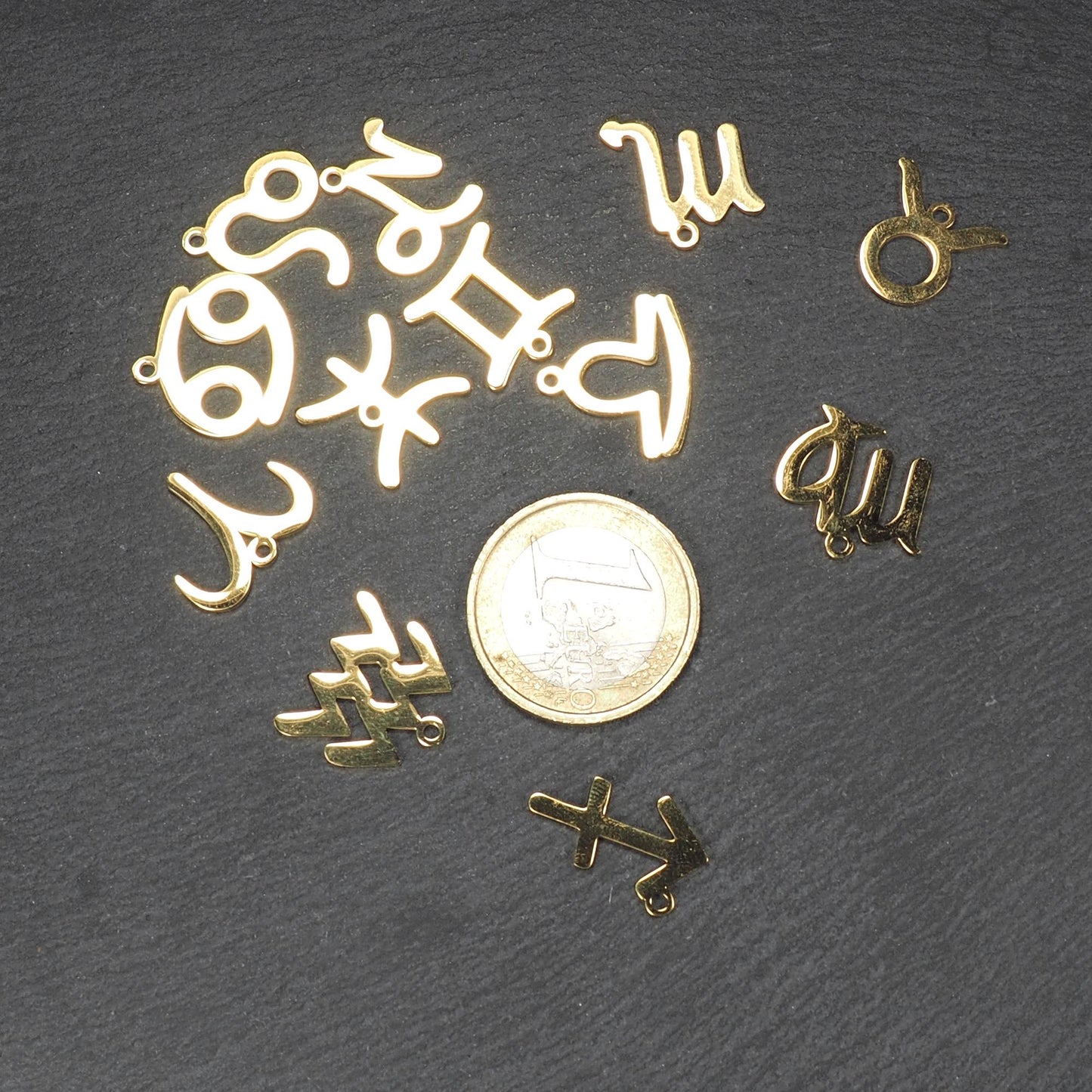 Mehrere flache, goldfarbene, glänzende Anhänger in durchbrochenen Tierkreiszeichenformen mit kleinen Ösen, neben einer 1-Euro-Münze.