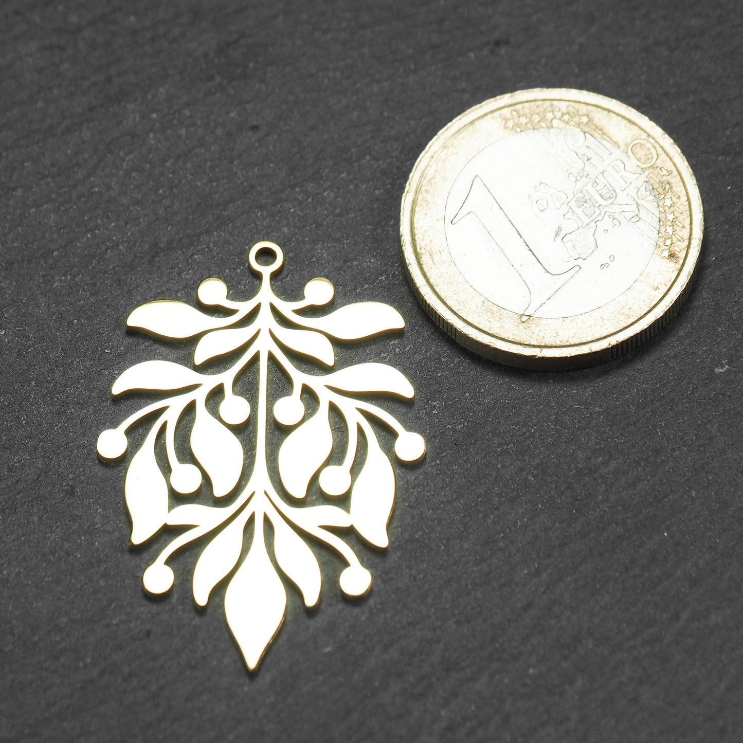 Goldfarbener Metallanhänger neben einer 1 Euro Münze zum Größenvergleich mit glänzender Oberfläche, durchbrochenes Zweig- und Beerenmotiv mit stilisierten Blättern und runder Öse oben.