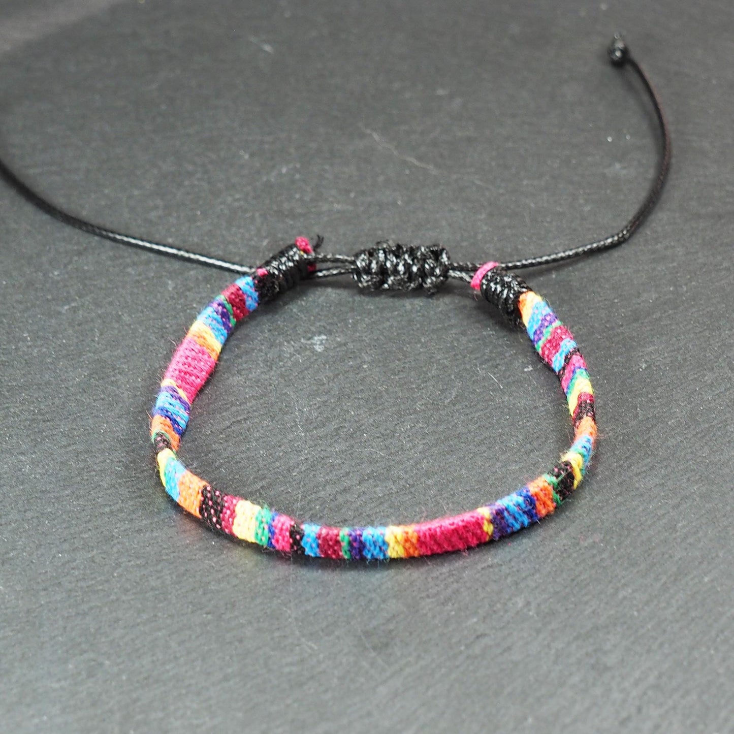 Gewebtes, flaches Armband mit regenbogenfarbenen Streifen, schmaler Form und feiner Webstruktur, schwarzer Schiebeknotenverschluss.