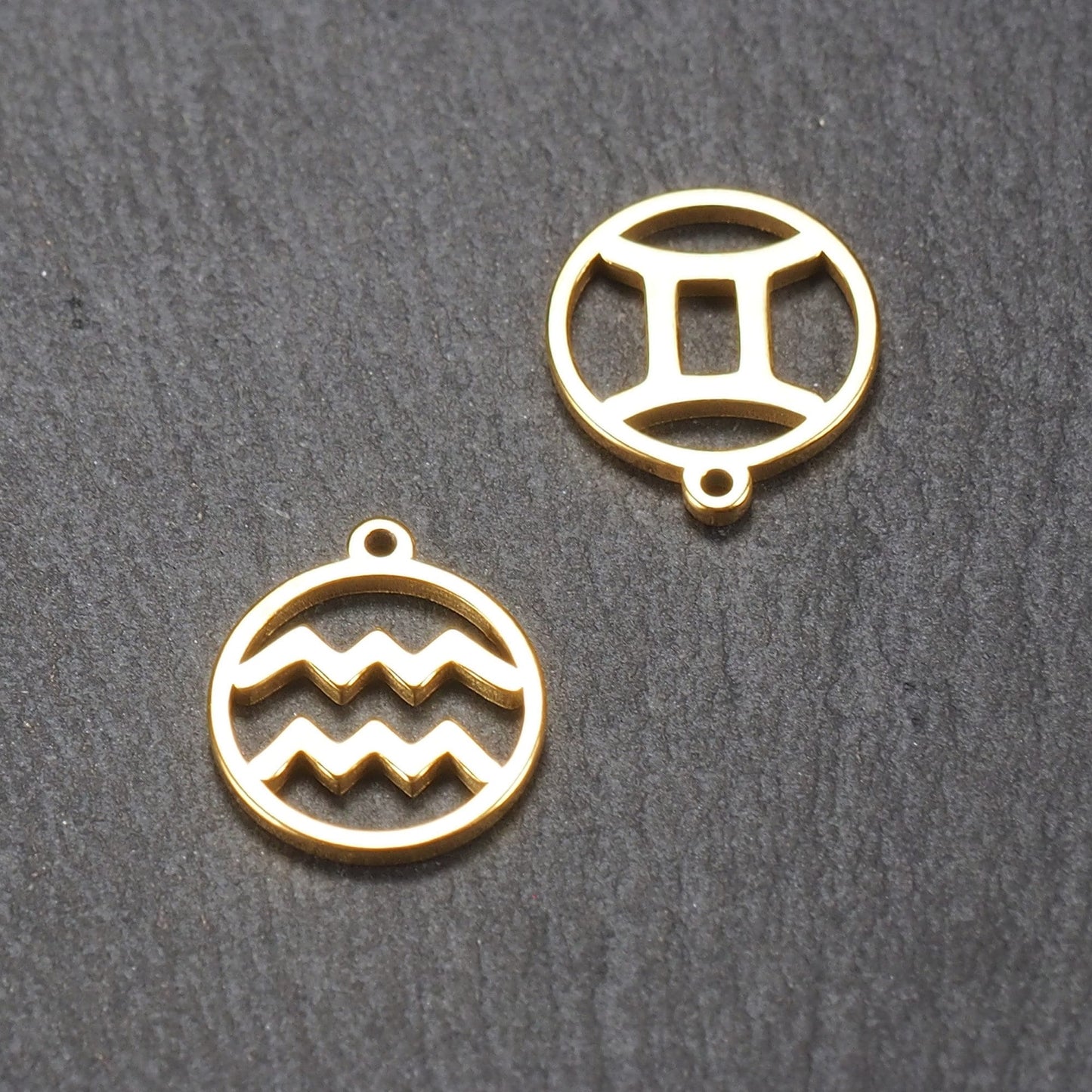 Zwei runde goldfarbene Anhänger mit glatter glänzender Oberfläche, filigranen Aussparungen (Wellen- und Zwillingsmotiv) und je einer integrierten Öse.