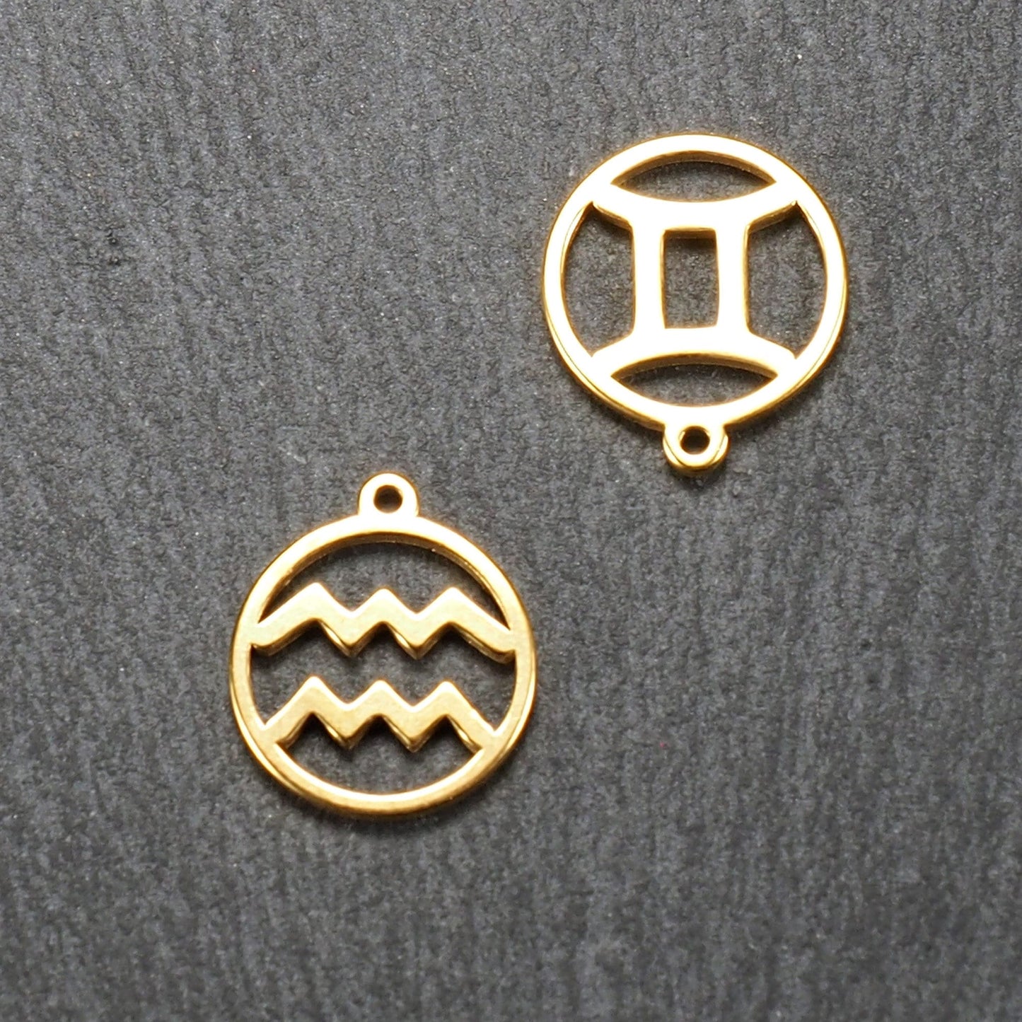 Zwei runde, goldfarbene, durchbrochene Anhänger mit glänzender Oberfläche; jeweils runder Rahmen mit Wellen- bzw. Zwillingsmotiv und kleiner Öse.