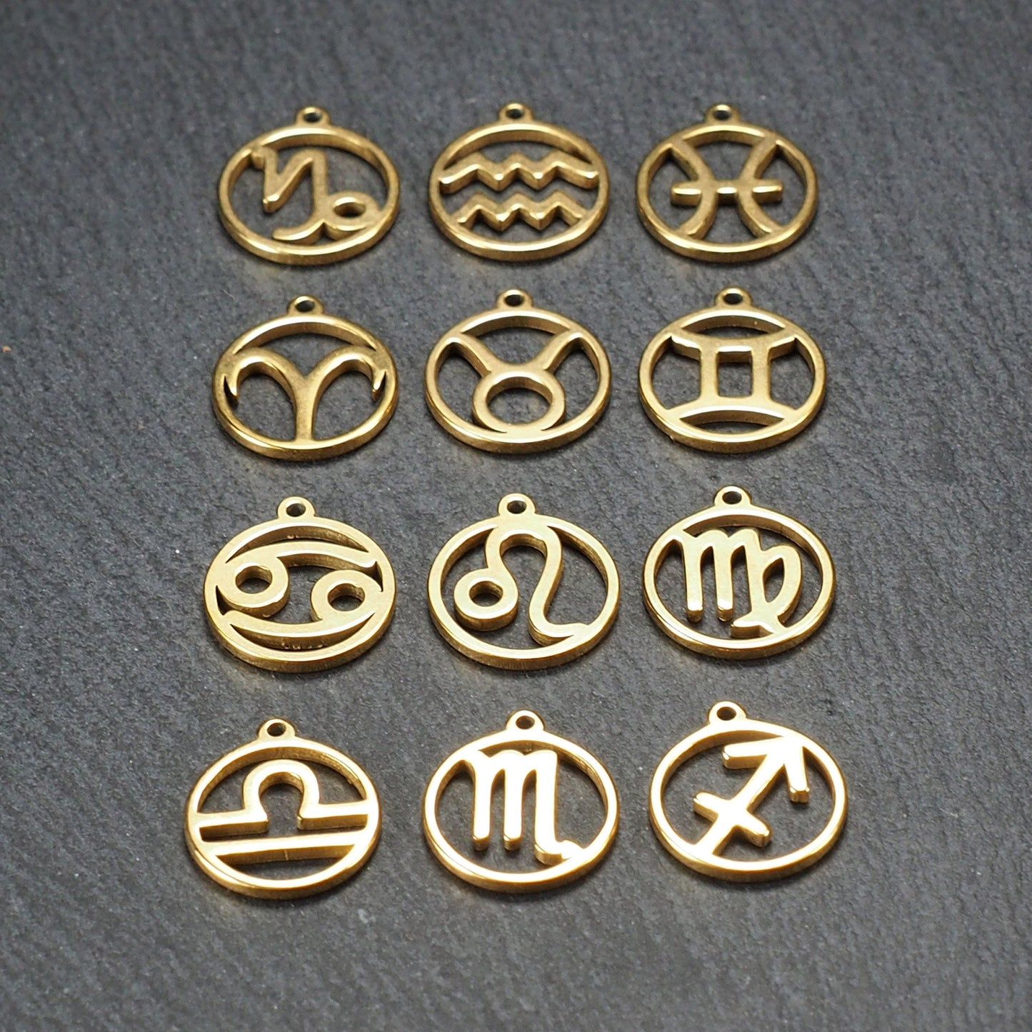Zwölf goldfarbene, glänzende, runde Anhänger mit durchbrochenen Sternzeichen-Symbolen in feinem Metallrahmen, jeweils mit kleiner Öse oben.