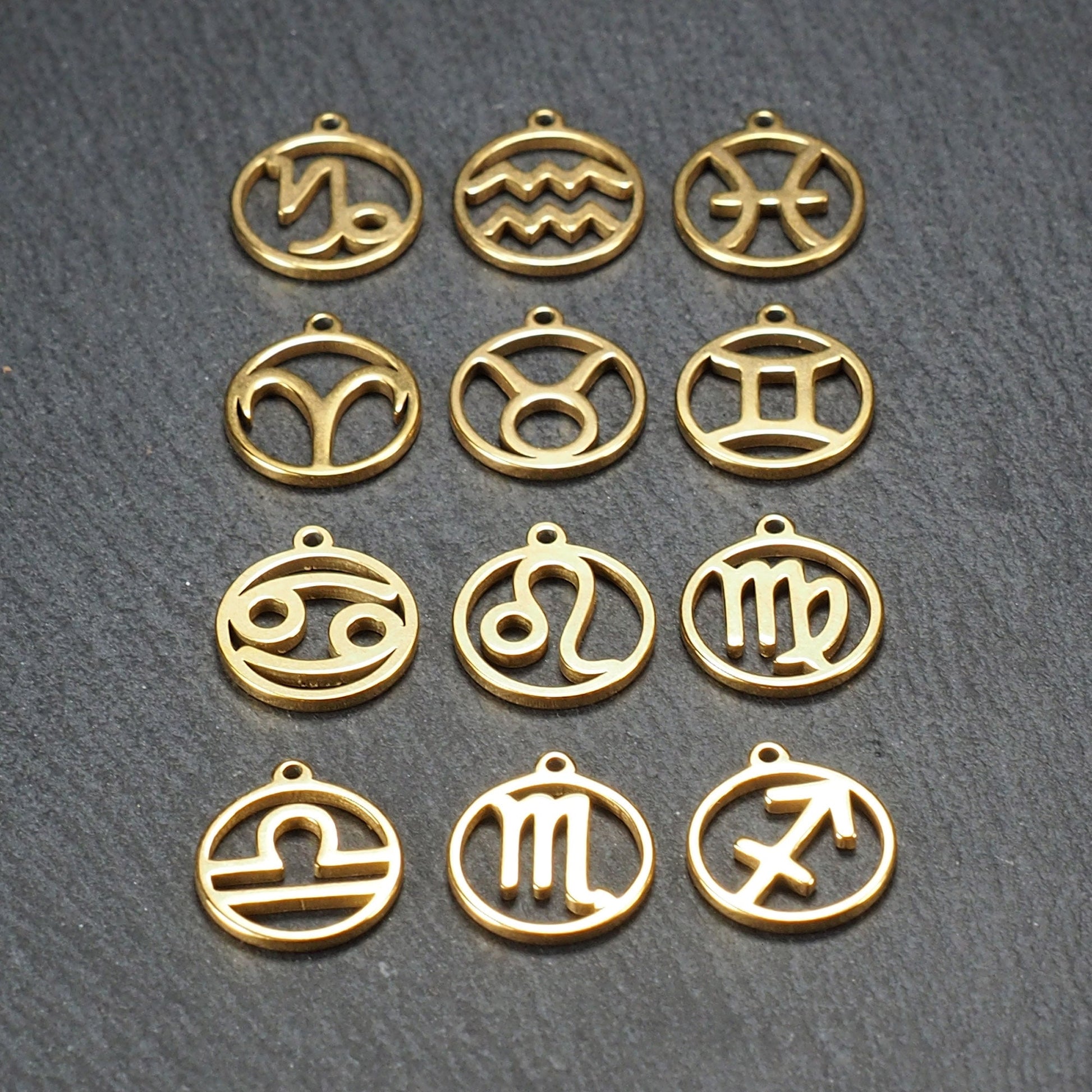 Zwölf goldfarbene, glänzende, runde Anhänger mit durchbrochenen Sternzeichen-Symbolen in feinem Metallrahmen, jeweils mit kleiner Öse oben.