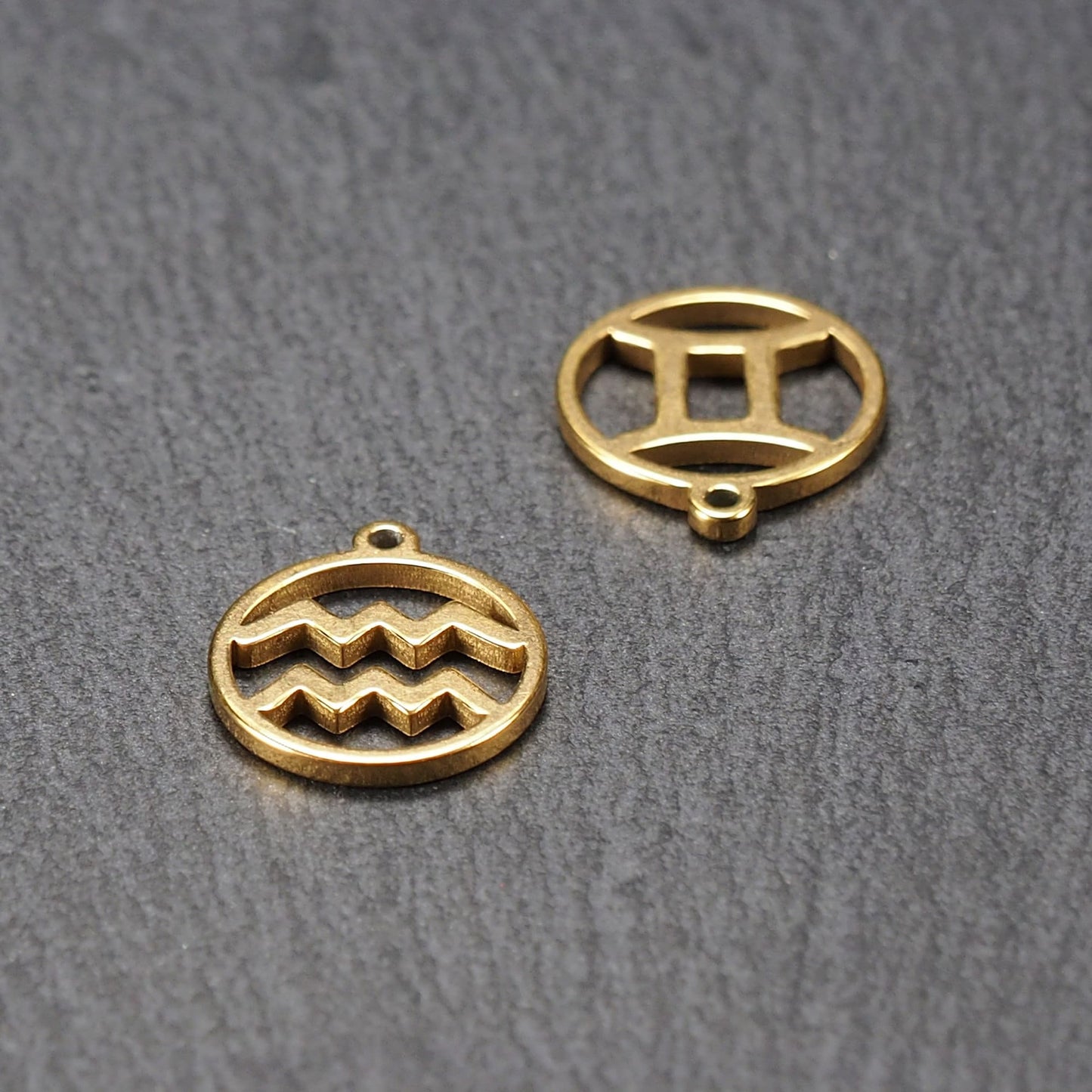 Runder, vergoldeter Metallanhänger, glatt glänzend, mit durchbrochenem Sternzeichen-Motiv (Wellenlinien und Zwillingssymbol) und integrierter Öse.