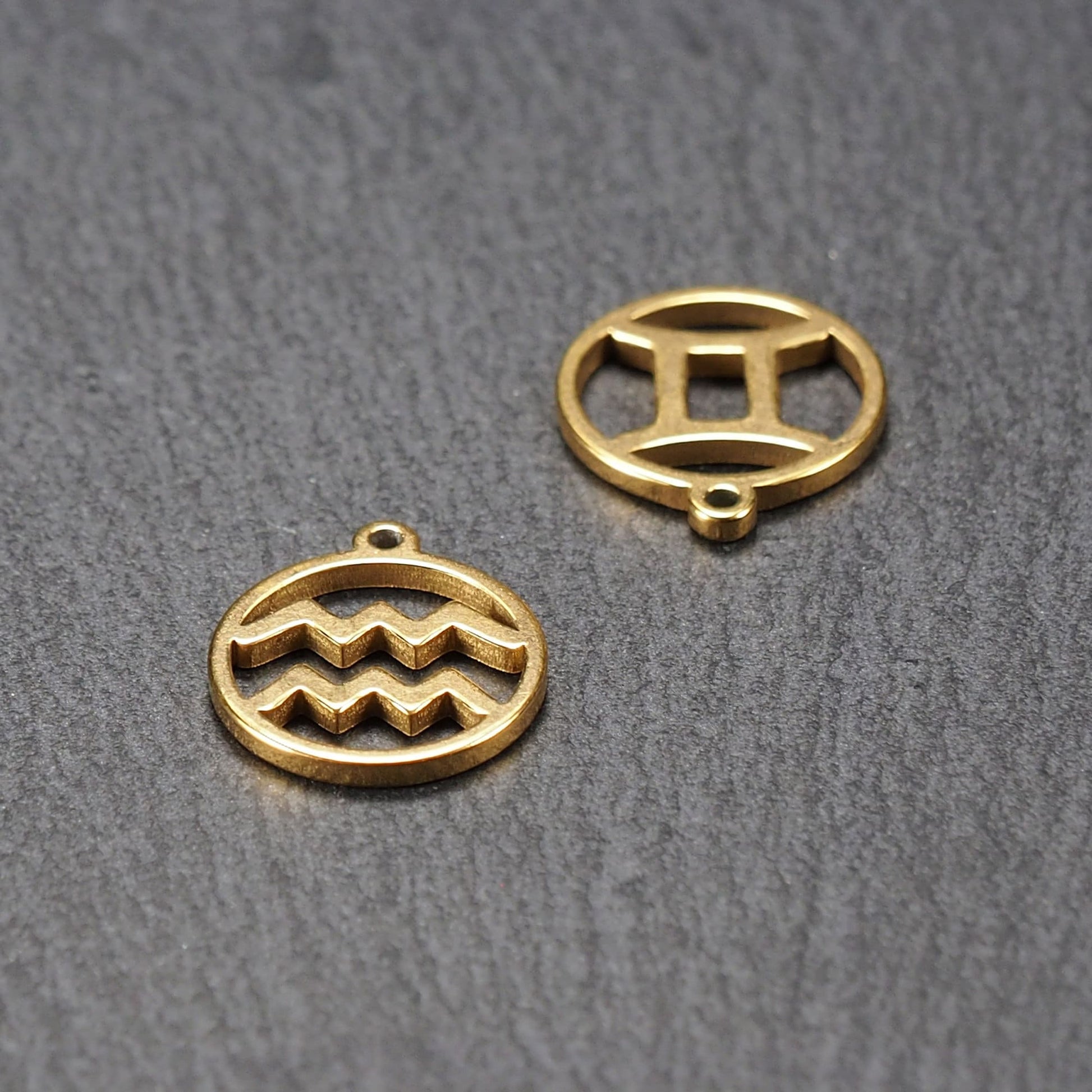 Runder, vergoldeter Metallanhänger, glatt glänzend, mit durchbrochenem Sternzeichen-Motiv (Wellenlinien und Zwillingssymbol) und integrierter Öse.