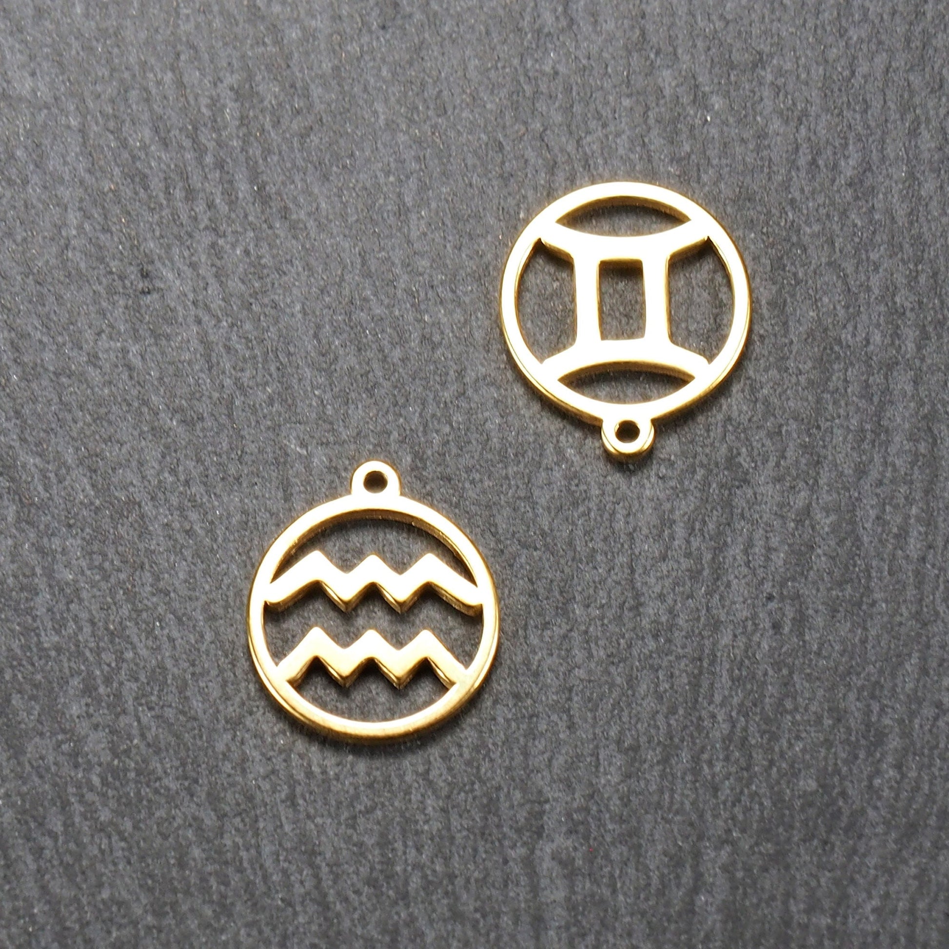 Zwei runde, vergoldete Anhänger mit durchbrochenen Sternzeichen-Motiven, glänzender glatter Oberfläche und integrierter Öse.