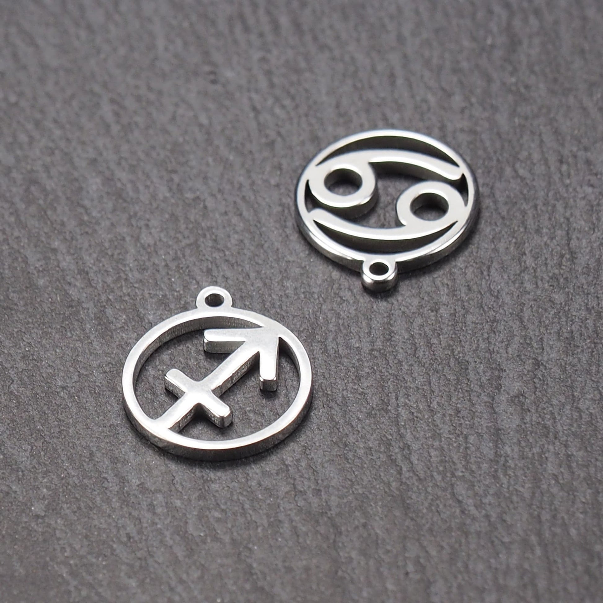 Zwei runde silberne Metallanhänger mit durchbrochenen Sternzeichensymbolen (Krebs, Schütze) und sichtbarer integrierter Öse auf dunklem Hintergrund.
