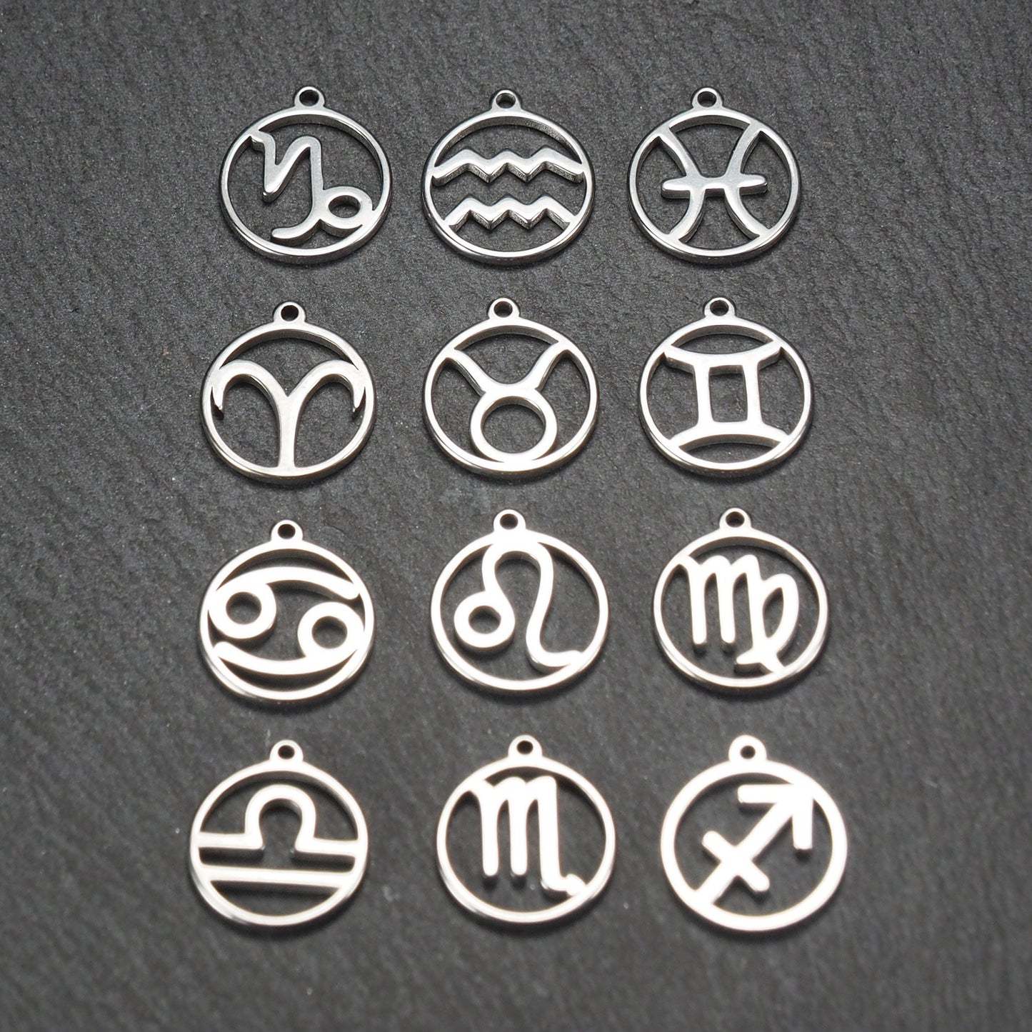 Zwölf runde silberne Anhänger mit durchbrochenen Sternzeichenmotiven, glänzende Oberfläche, integrierte Öse, auf dunklem strukturiertem Untergrund.
