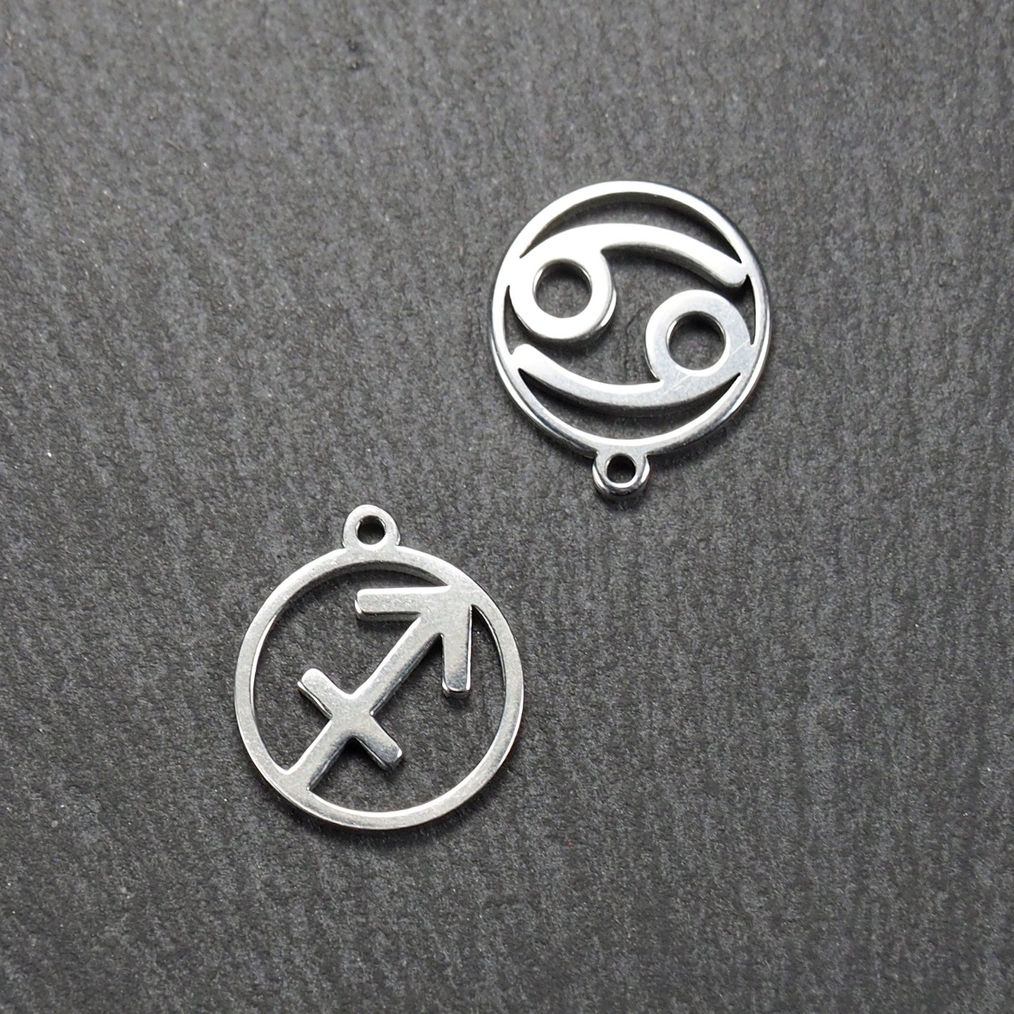 Zwei runde silberne Metallanhänger mit durchbrochenen Sternzeichensymbolen (Krebs, Schütze) und sichtbarer integrierter Öse auf dunklem Hintergrund.