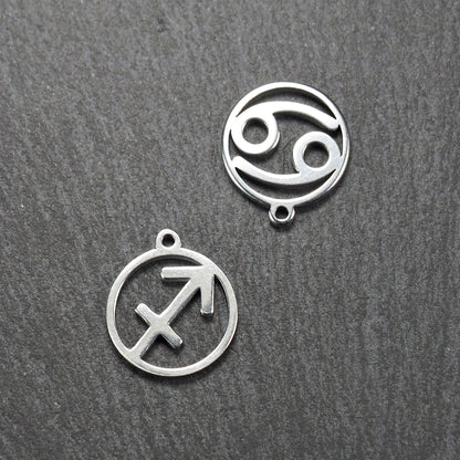 Zwei runde silberne Metallanhänger mit durchbrochenen Sternzeichensymbolen (Krebs, Schütze) und sichtbarer integrierter Öse auf dunklem Hintergrund.