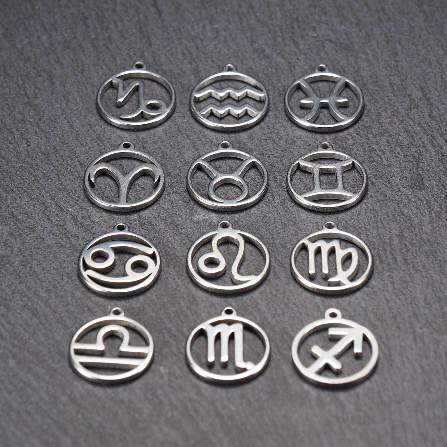 Zwölf runde silberne Anhänger mit durchbrochenen Sternzeichenmotiven, glänzende Oberfläche, integrierte Öse, auf dunklem strukturiertem Untergrund.