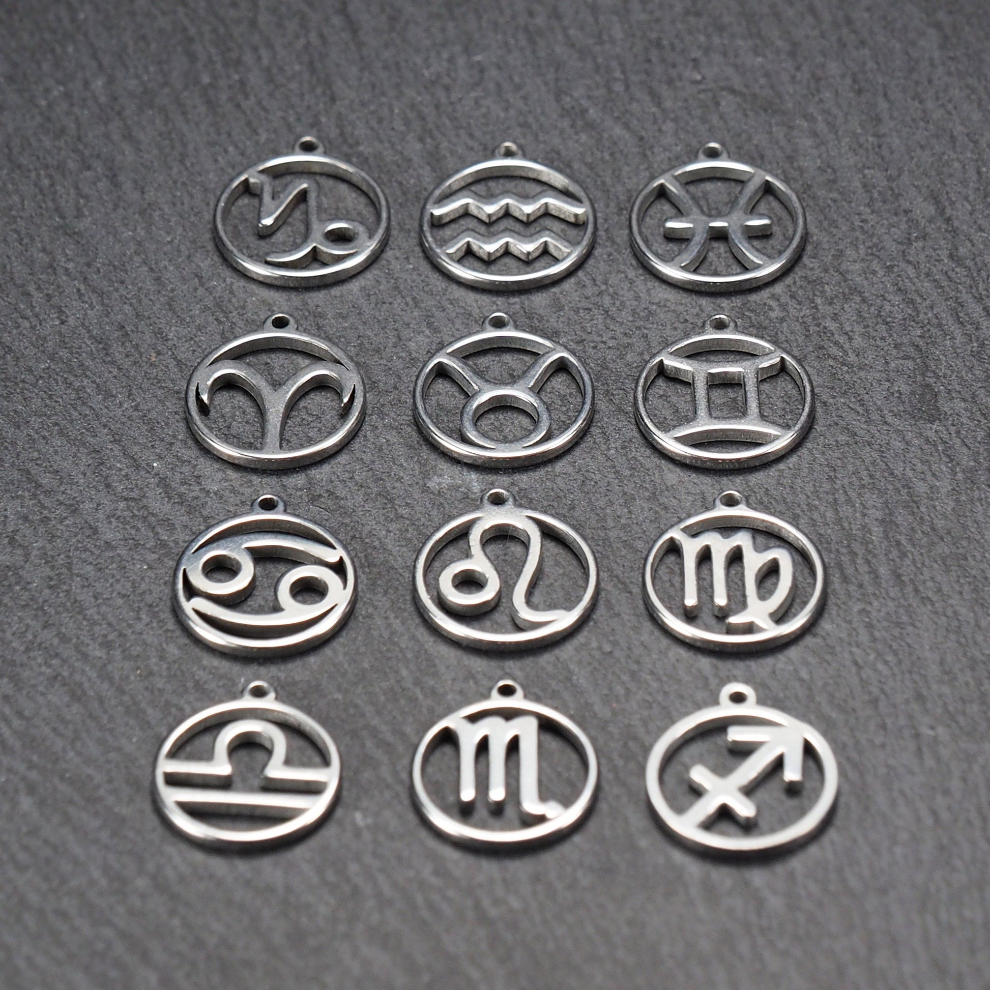 Zwölf runde silberne Anhänger mit durchbrochenen Sternzeichenmotiven, glänzende Oberfläche, integrierte Öse, auf dunklem strukturiertem Untergrund.