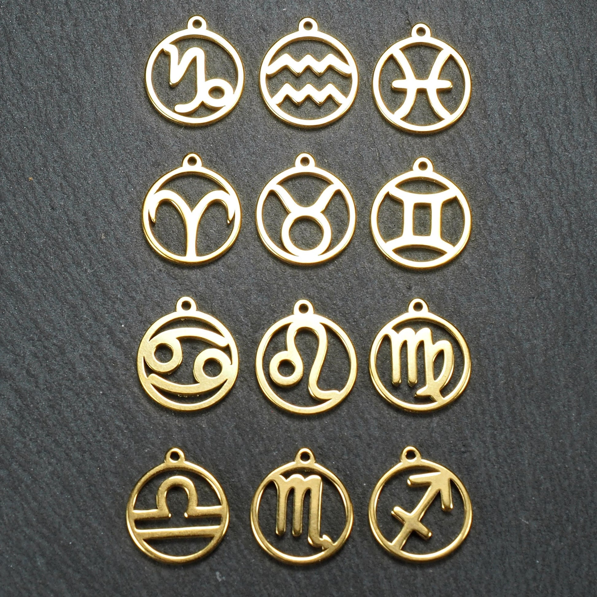 Zwölf goldfarbene, glänzende, runde Anhänger mit durchbrochenen Sternzeichen-Symbolen in feinem Metallrahmen, jeweils mit kleiner Öse oben.