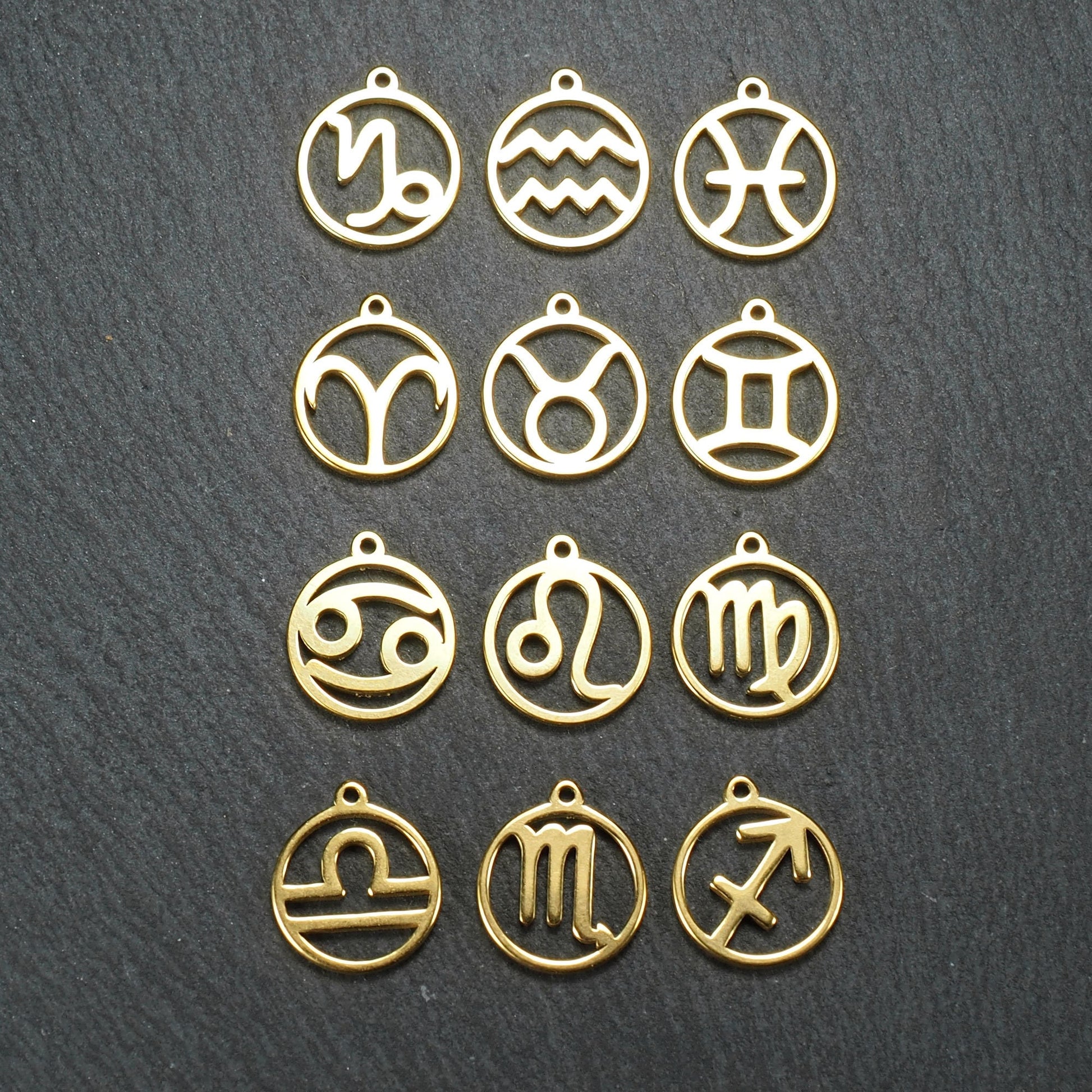 Zwölf goldfarbene, glänzende, runde Anhänger mit durchbrochenen Sternzeichen-Symbolen in feinem Metallrahmen, jeweils mit kleiner Öse oben.