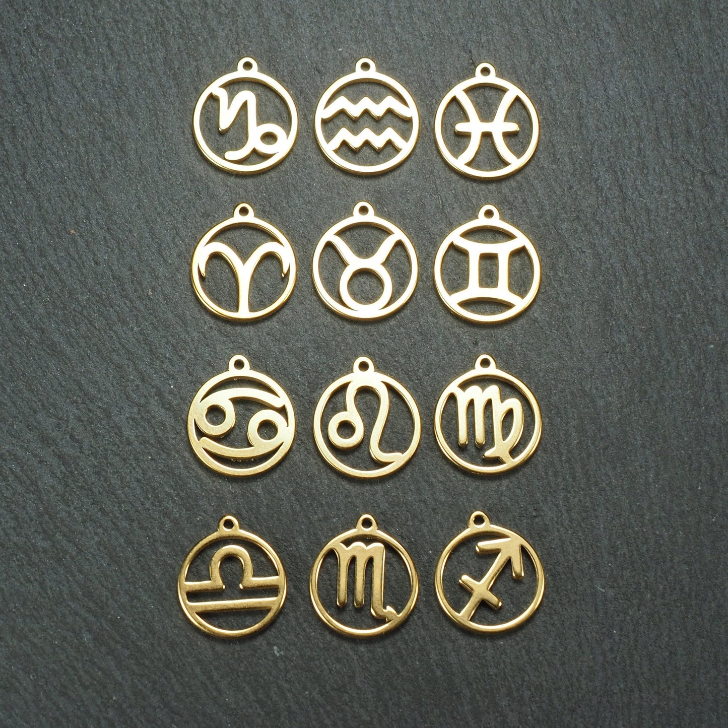 Zwölf goldfarbene, glänzende, runde Anhänger mit durchbrochenen Sternzeichen-Symbolen in feinem Metallrahmen, jeweils mit kleiner Öse oben.