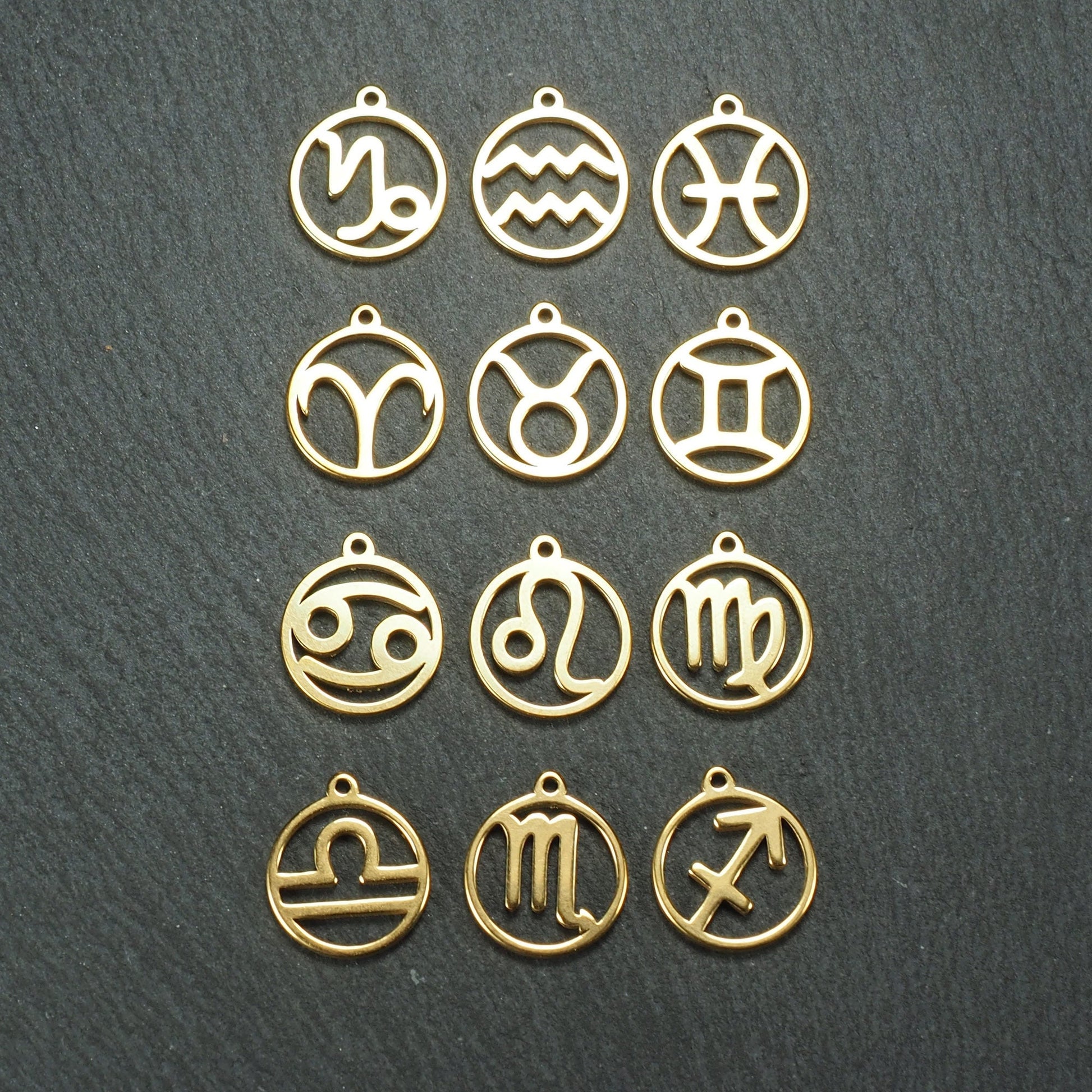 Zwölf goldfarbene, glänzende, runde Anhänger mit durchbrochenen Sternzeichen-Symbolen in feinem Metallrahmen, jeweils mit kleiner Öse oben.