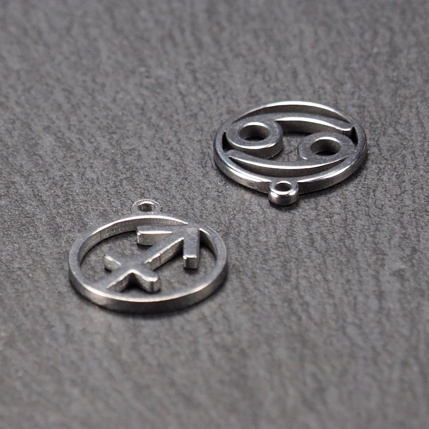Zwei runde silberne Metallanhänger mit durchbrochenen Sternzeichensymbolen (Krebs, Schütze) und sichtbarer integrierter Öse auf dunklem Hintergrund.