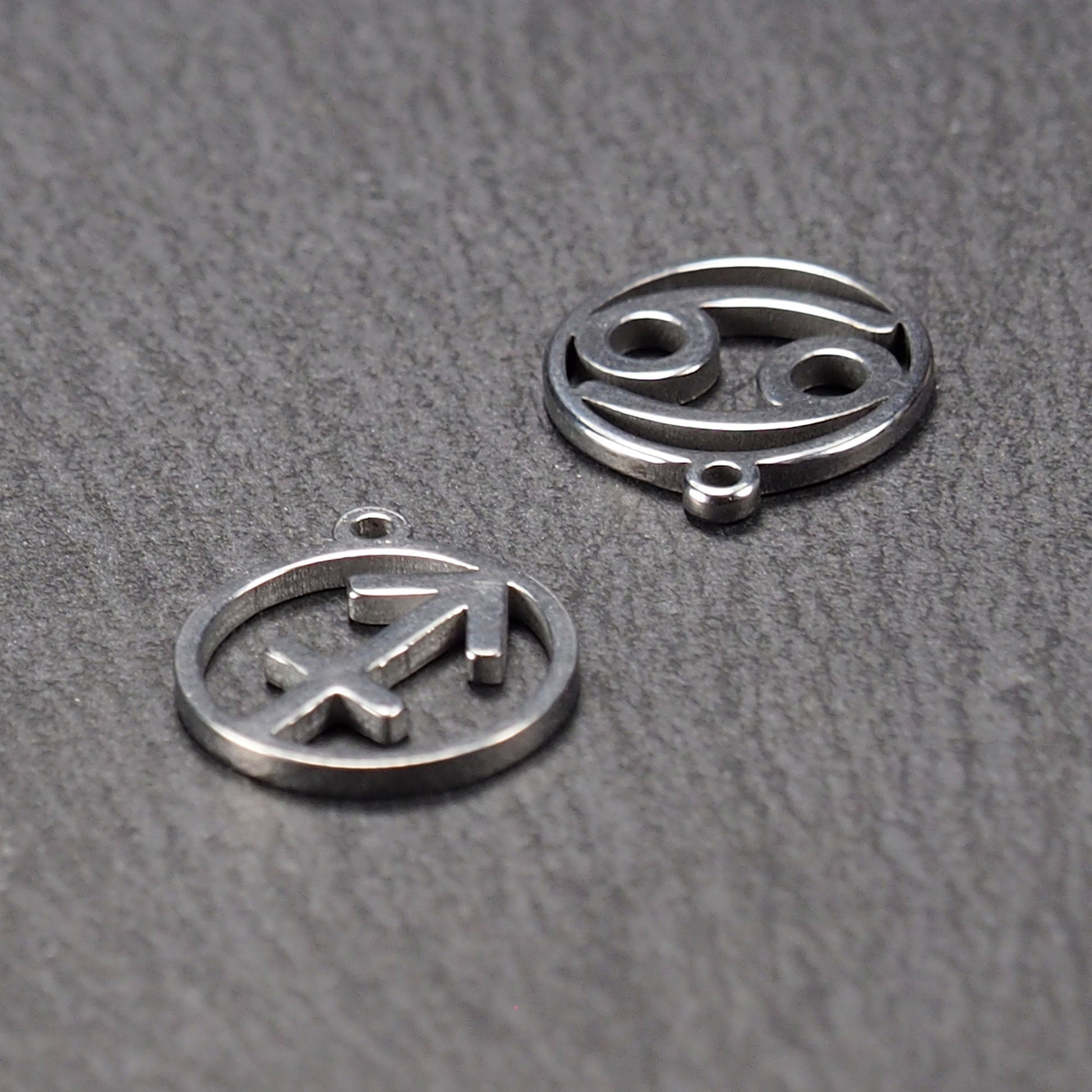 Zwei runde silberne Metallanhänger mit durchbrochenen Sternzeichensymbolen (Krebs, Schütze) und sichtbarer integrierter Öse auf dunklem Hintergrund.