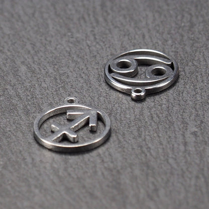 Zwei runde silberne Metallanhänger mit durchbrochenen Sternzeichensymbolen (Krebs, Schütze) und sichtbarer integrierter Öse auf dunklem Hintergrund.
