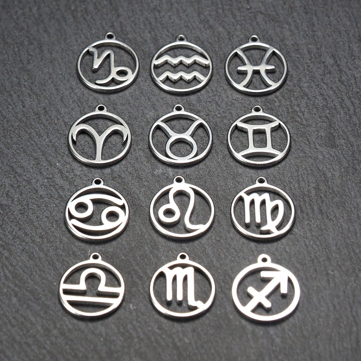 Zwölf runde silberne Anhänger mit durchbrochenen Sternzeichenmotiven, glänzende Oberfläche, integrierte Öse, auf dunklem strukturiertem Untergrund.