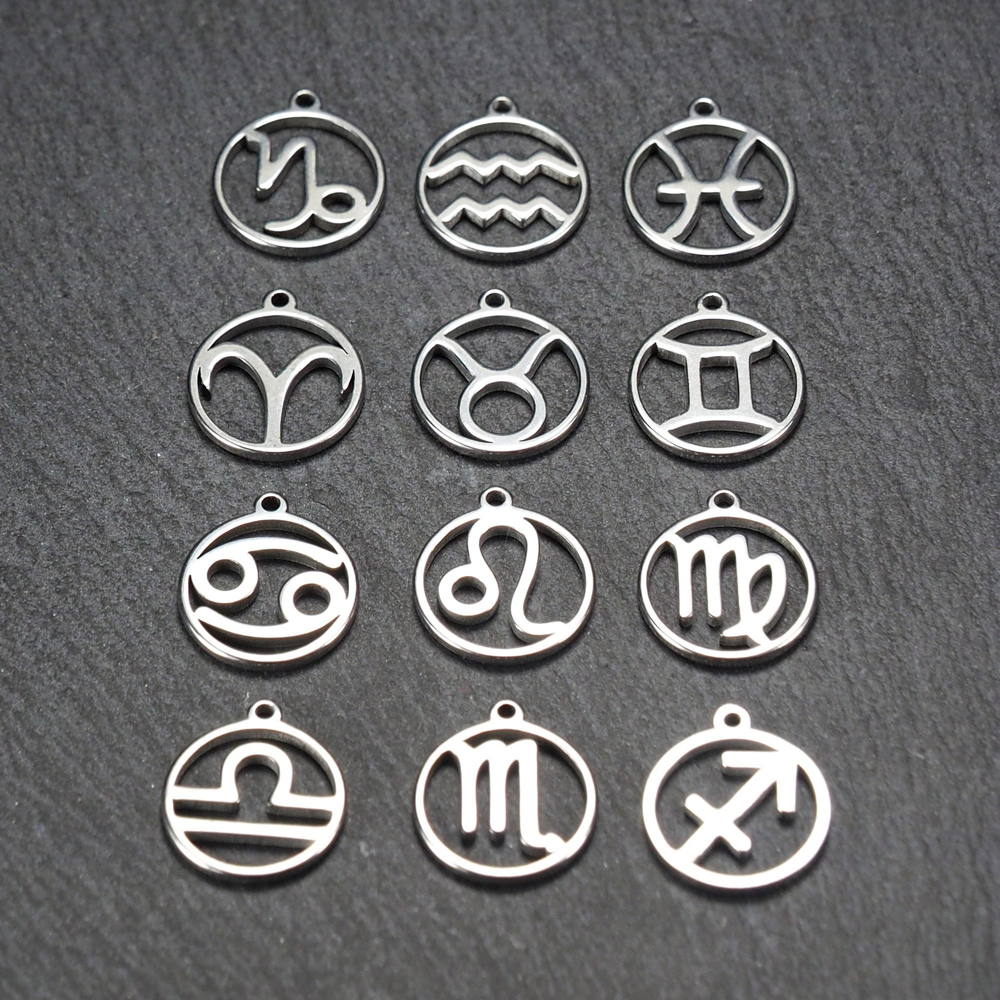 Zwölf runde silberne Anhänger mit durchbrochenen Sternzeichenmotiven, glänzende Oberfläche, integrierte Öse, auf dunklem strukturiertem Untergrund.