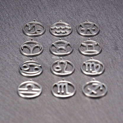 Zwölf runde silberne Anhänger mit durchbrochenen Sternzeichenmotiven, glänzende Oberfläche, integrierte Öse, auf dunklem strukturiertem Untergrund.