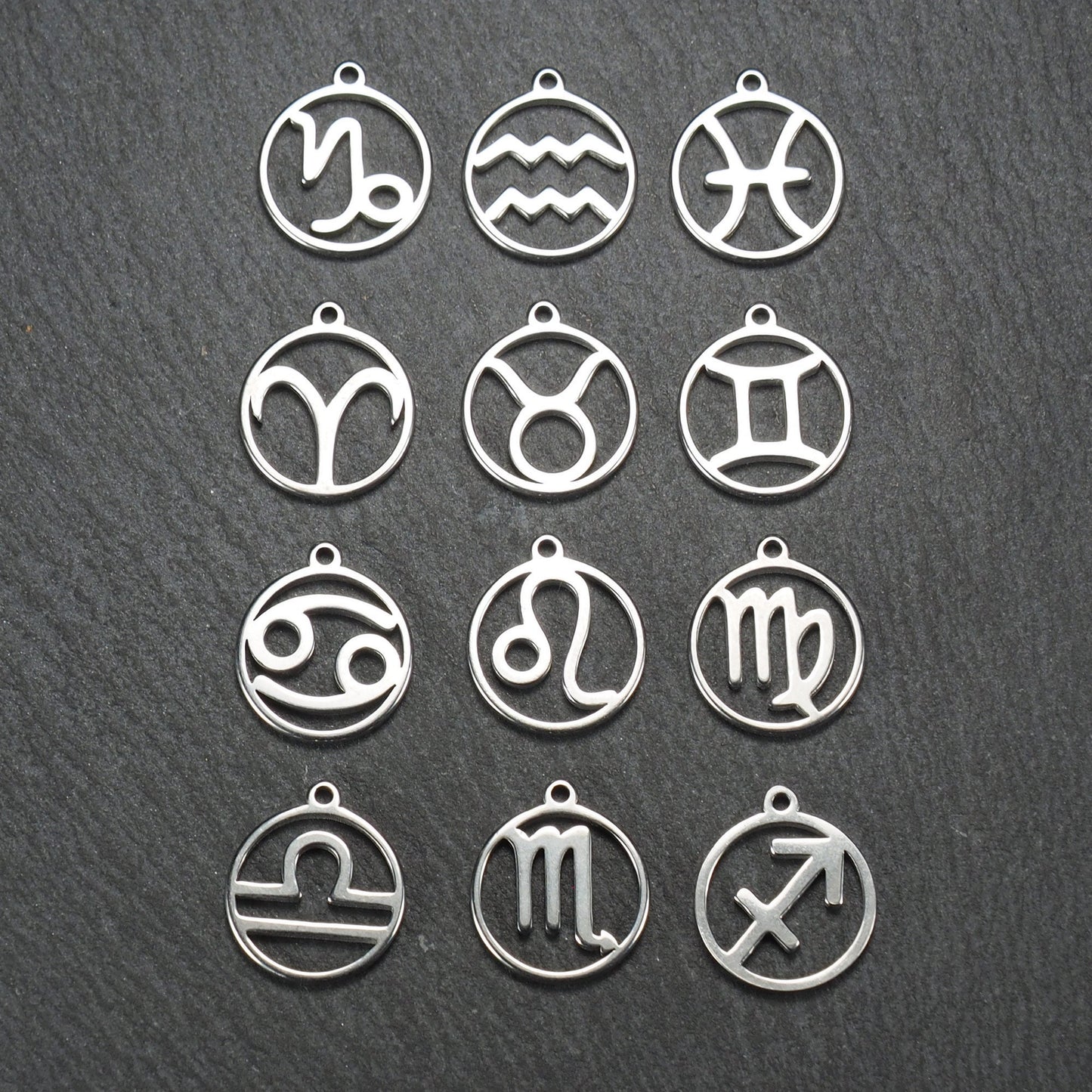 Zwölf runde silberne Anhänger mit durchbrochenen Sternzeichenmotiven, glänzende Oberfläche, integrierte Öse, auf dunklem strukturiertem Untergrund.