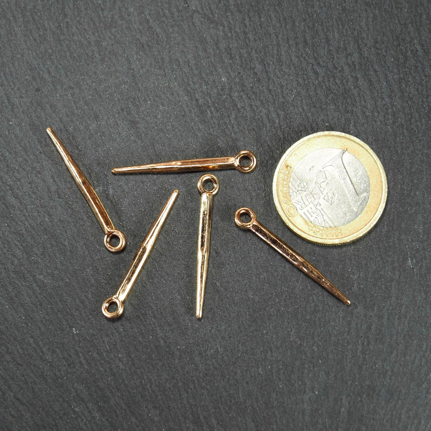 Fünf goldfarbene, konische Anhänger mit sichtbarer Öse und glänzender Oberfläche, spitz zulaufend, neben einer 1-Euro-Münze.