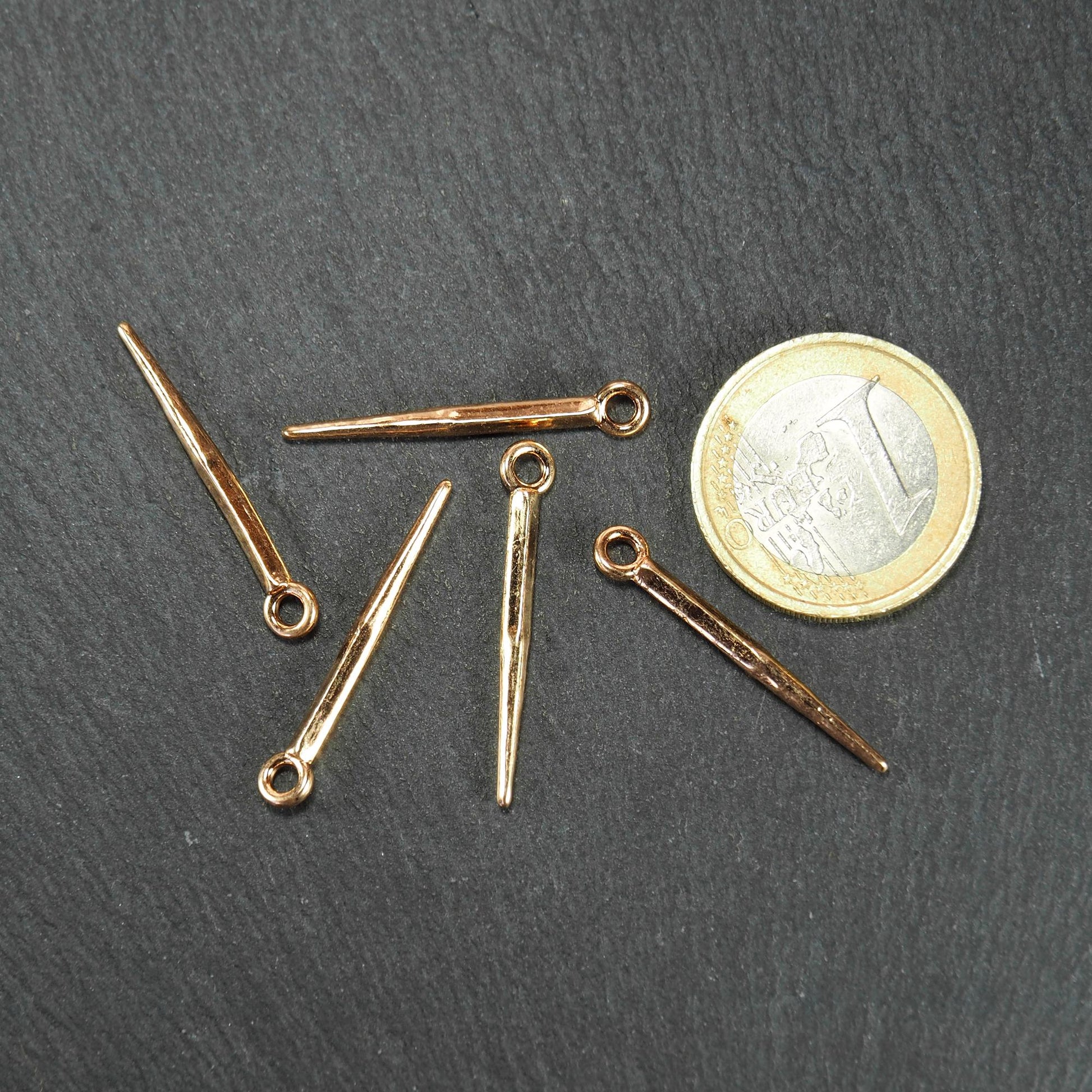 Fünf goldfarbene, konische Anhänger mit sichtbarer Öse und glänzender Oberfläche, spitz zulaufend, neben einer 1-Euro-Münze.
