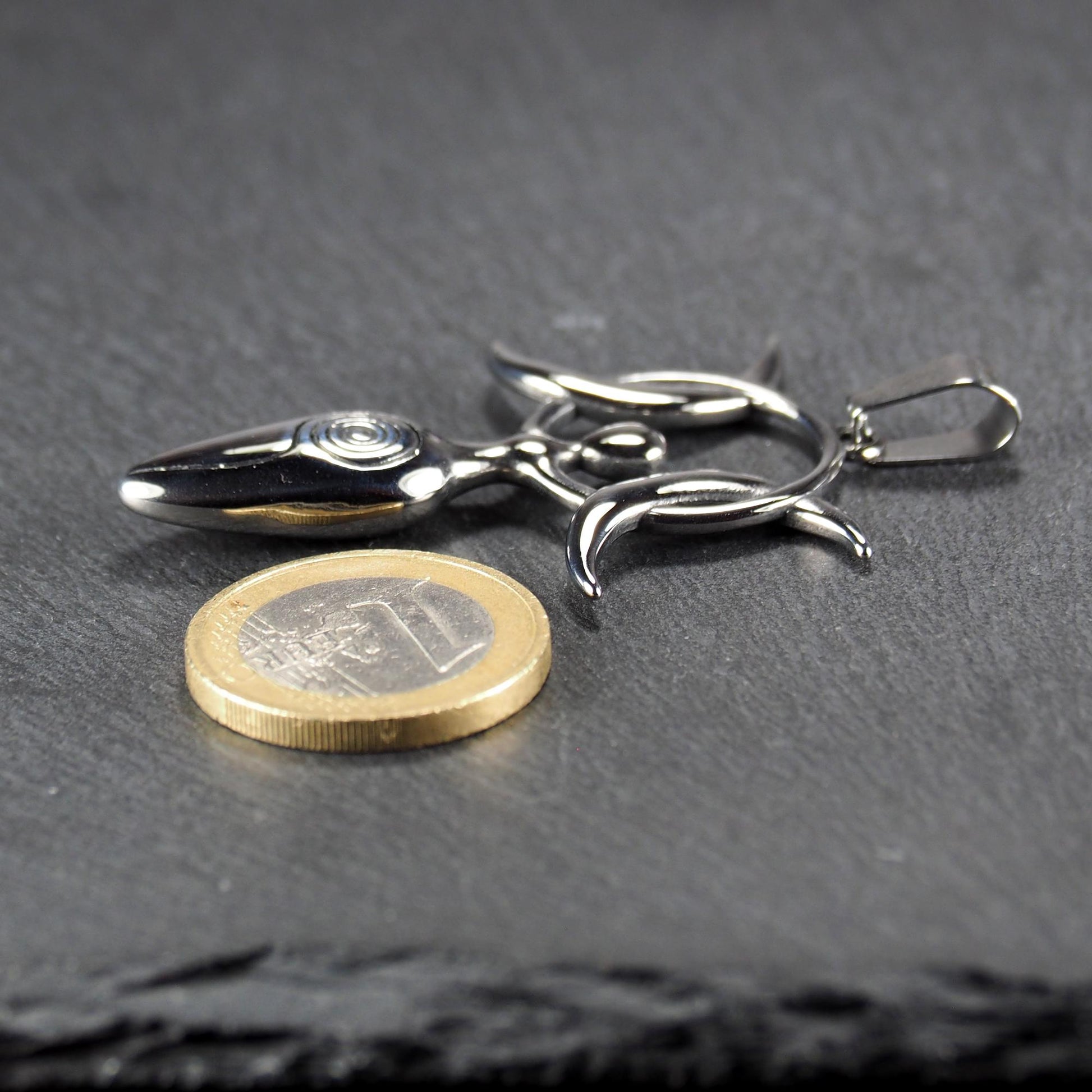 Figürlicher silberner, polierter Anhänger mit durchbrochenem Kreis, drei Mondsicheln, ovalem Kopf mit Spirale und Aufhängeöse; Ein-Euro-Münze als Größenvergleich.