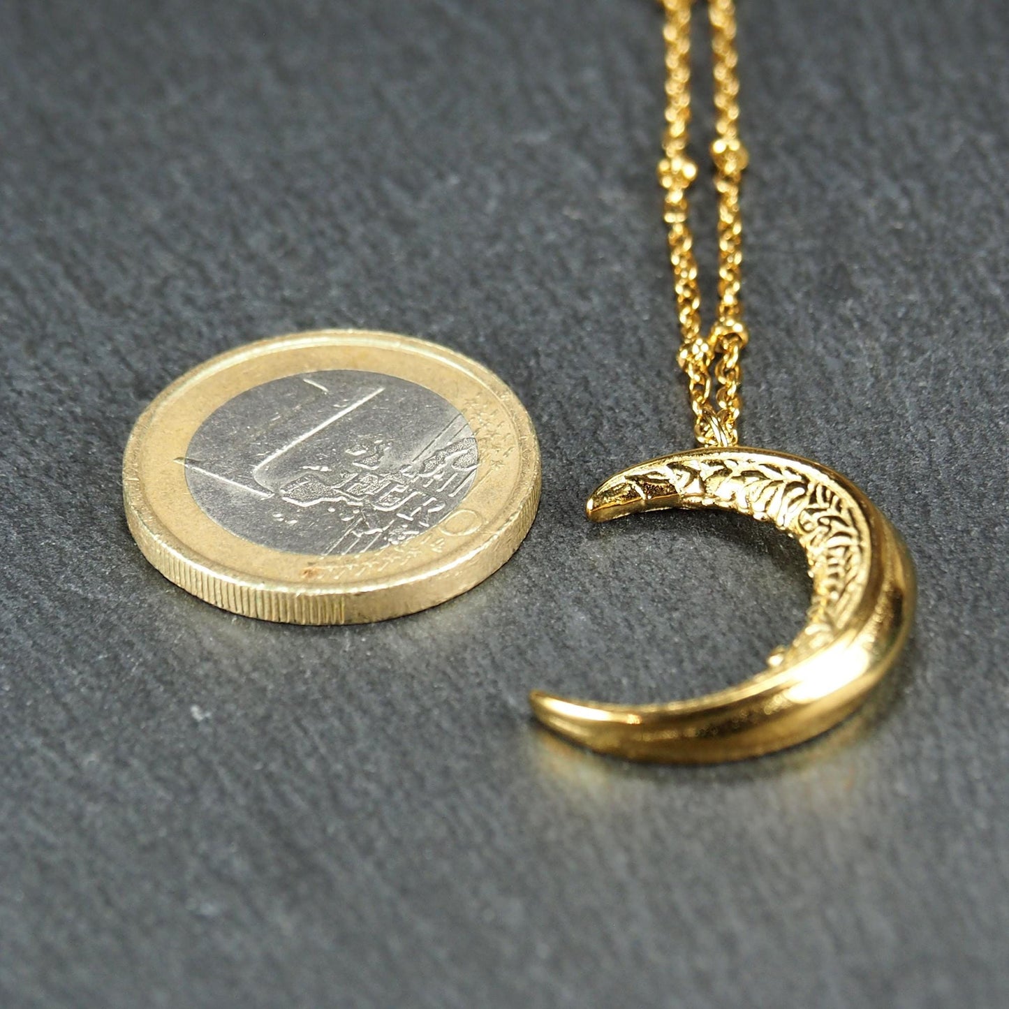 Mondsichel-Anhänger, goldfarbene Oberfläche mit filigraner Prägung an feiner Kette neben einer Euromünze.