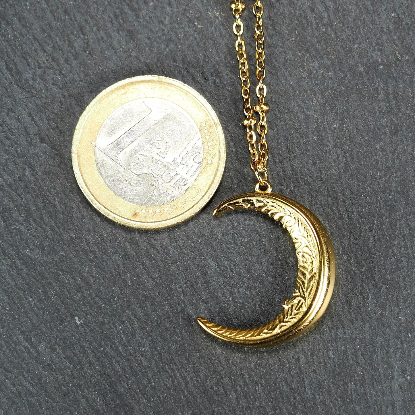 Mondsichel-Anhänger, goldfarbene Oberfläche mit filigraner Prägung an feiner Kette neben einer Euromünze.