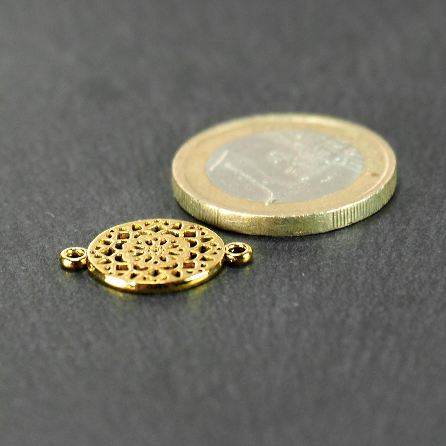 Runder goldfarbener Verbinder mit radialem Mandala-Blütenmuster und zwei Ösen; daneben 1-Euro-Münze als Größenvergleich.