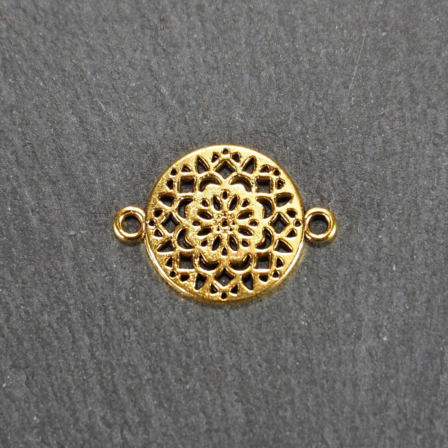 Runder goldfarbener Verbinder mit durchbrochenem, radial-symmetrischem Mandala-/Blütenmuster und zwei gegenüberliegenden Ösen.