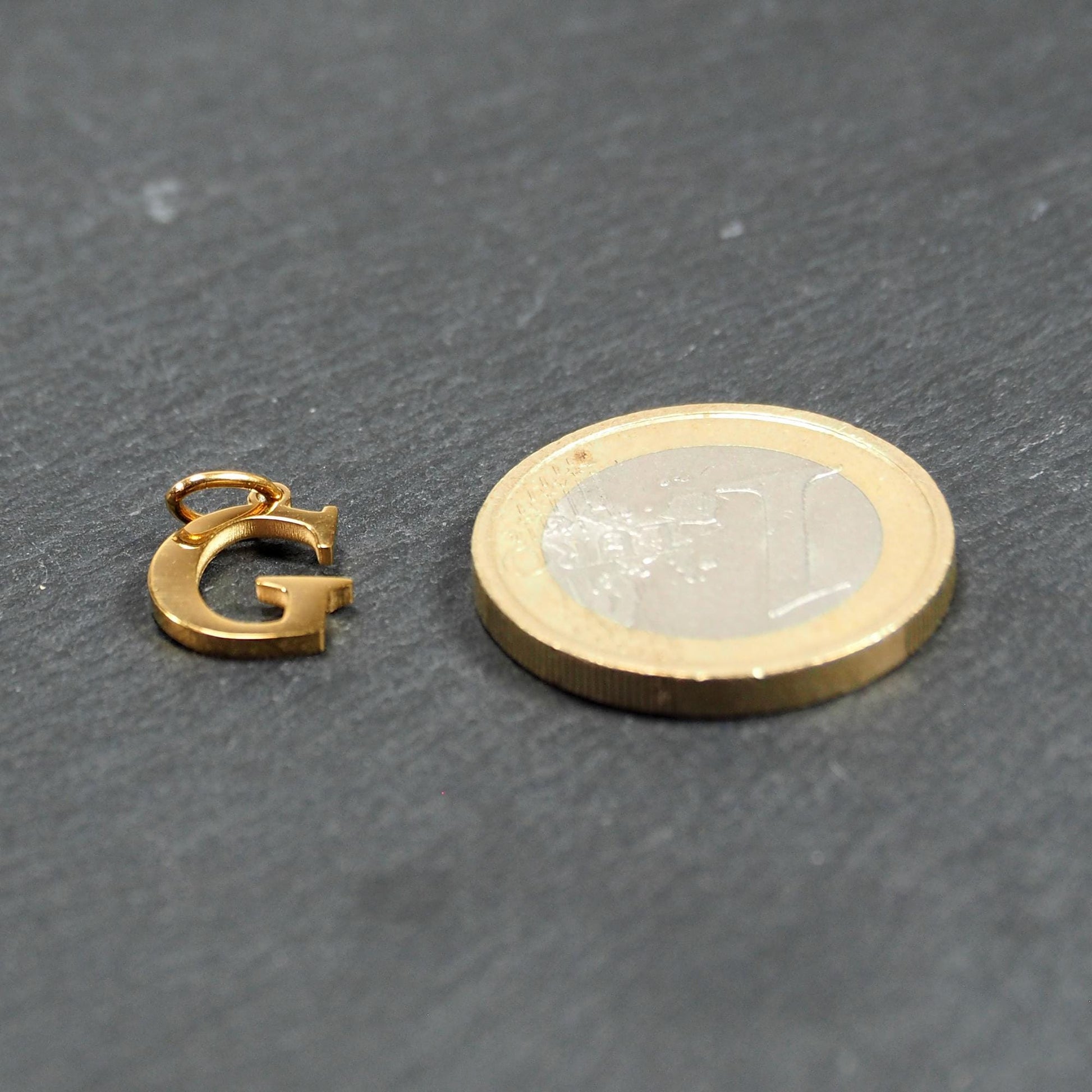 Goldfarbener, glänzender Buchstabenanhänger in Versal-Serifenschrift (Großbuchstabe G) mit kleiner Öse, neben einer 1-Euro-Münze zur Größendarstellung.