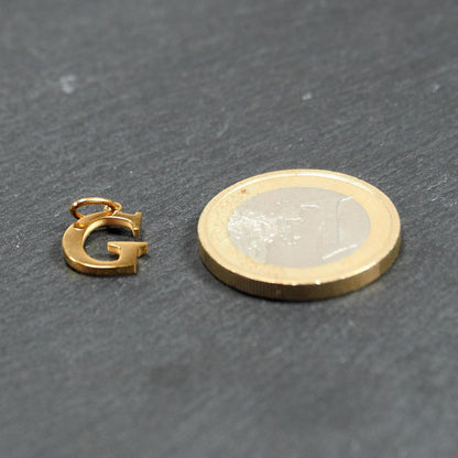 Goldfarbener, glänzender Buchstabenanhänger in Versal-Serifenschrift (Großbuchstabe G) mit kleiner Öse, neben einer 1-Euro-Münze zur Größendarstellung.