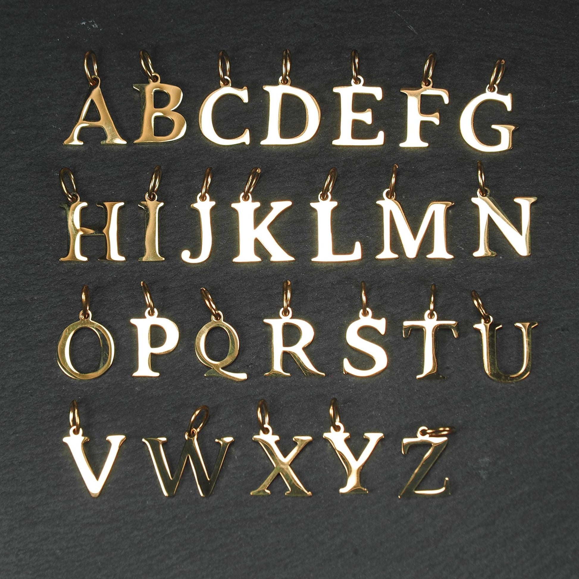 Goldfarbene, glänzende Buchstabenanhänger in Versal-Serifen-Schrift, einzelne Großbuchstaben A–Z mit jeweils kleiner Öse oben auf dunklem Hintergrund.
