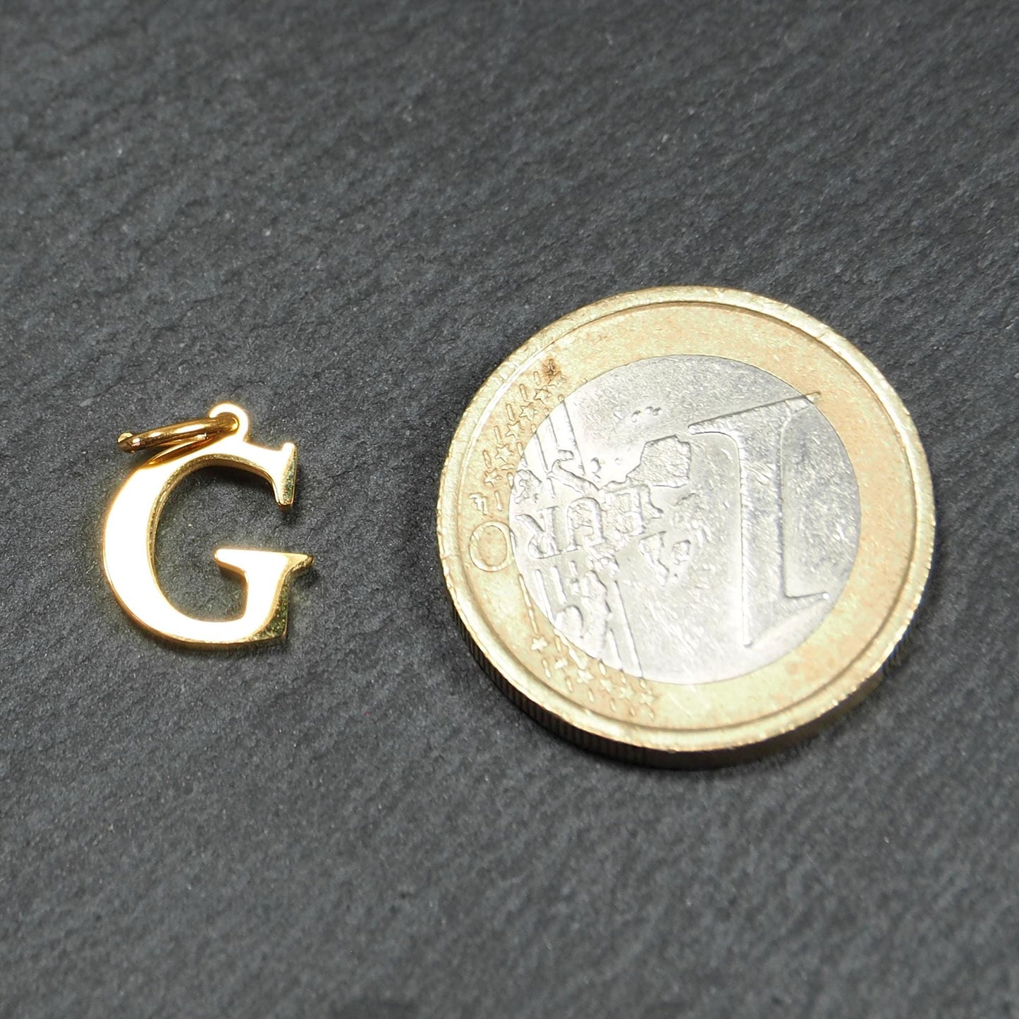 Goldfarbener, glänzender Buchstabenanhänger in Versal-Serifenschrift (Großbuchstabe G) mit kleiner Öse, neben einer 1-Euro-Münze zur Größendarstellung.