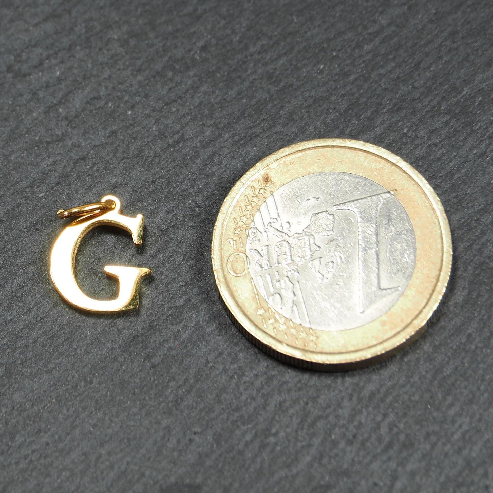 Goldfarbener, glänzender Buchstabenanhänger in Versal-Serifenschrift (Großbuchstabe G) mit kleiner Öse, neben einer 1-Euro-Münze zur Größendarstellung.