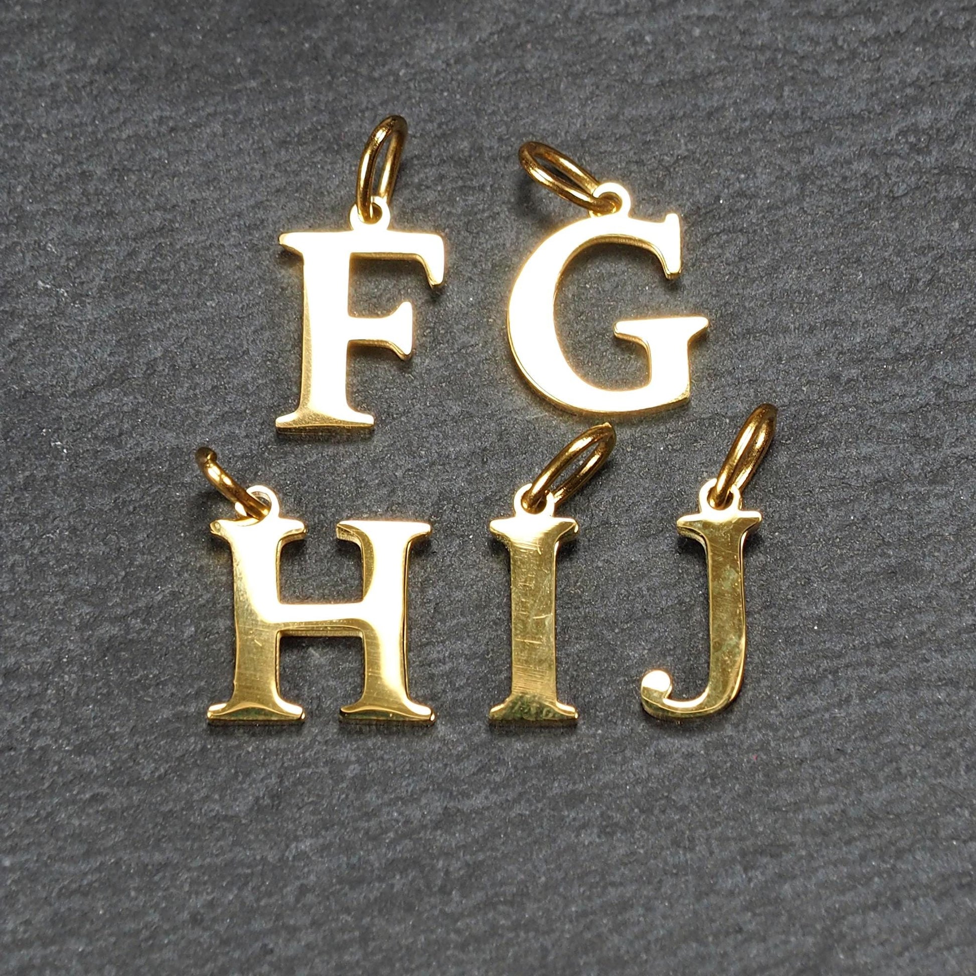 Fünf goldfarbene, glänzende Großbuchstaben (F, G, H, I, J) als serifenbetonte Anhänger mit kleinen Ösen.