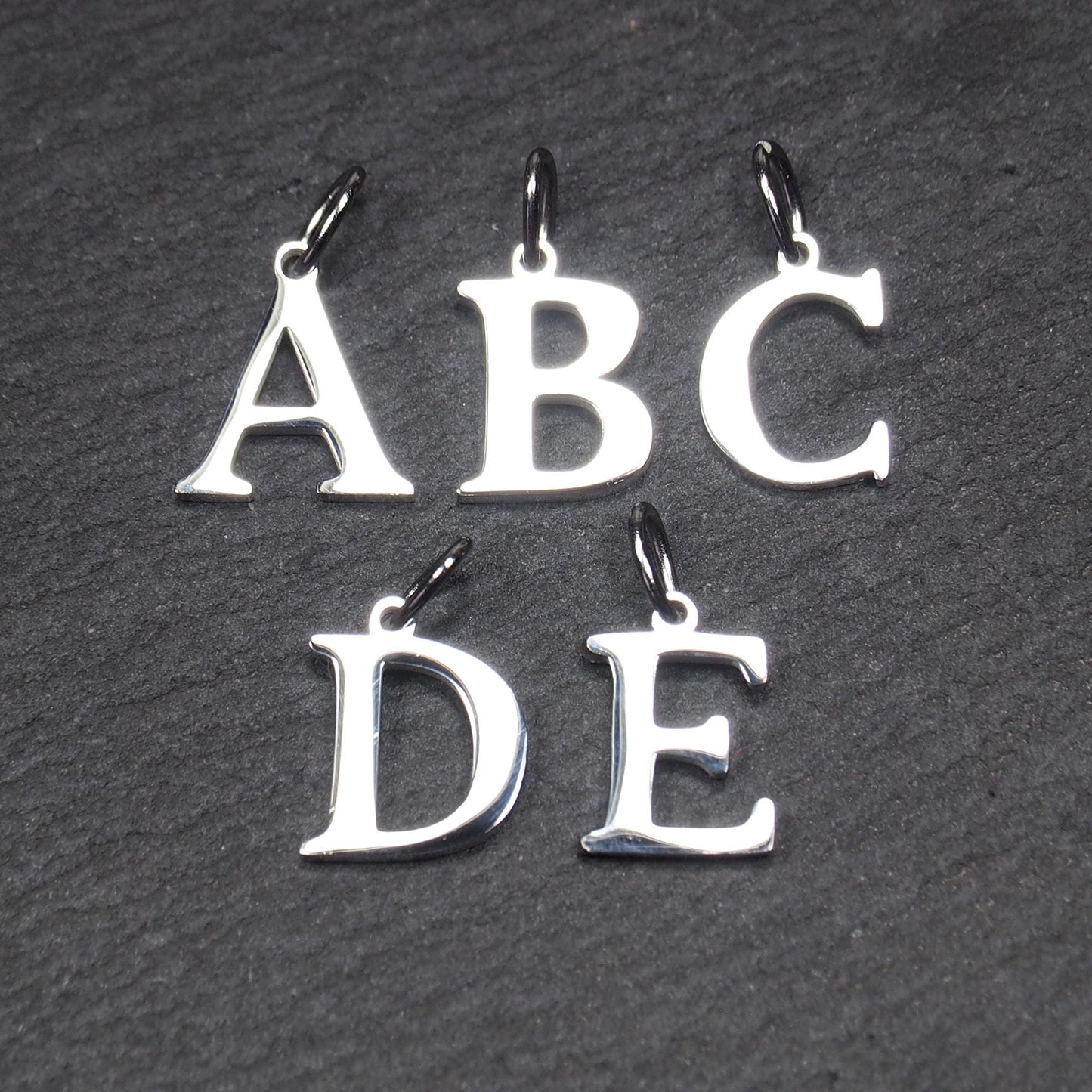 Fünf polierte silberne Großbuchstaben-Anhänger (A, B, C, D, E) in serifen-Typografie mit kleinen Ösen auf dunklem strukturiertem Hintergrund.