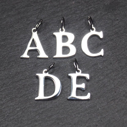 Fünf polierte silberne Großbuchstaben-Anhänger (A, B, C, D, E) in serifen-Typografie mit kleinen Ösen auf dunklem strukturiertem Hintergrund.