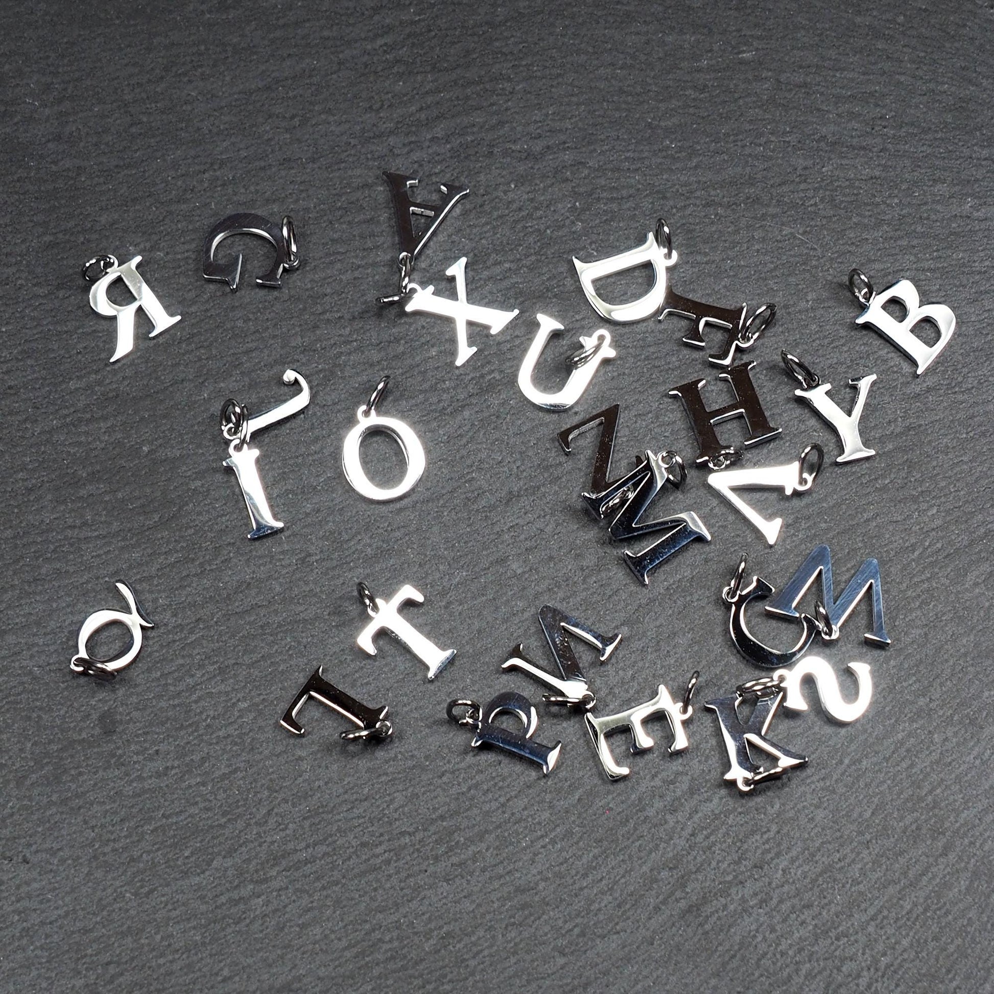 Buchstaben-Anhänger, silberne polierte Metalloberfläche, Großbuchstaben in Serifenschrift, kompakte typografische Form, jeweils mit kleiner Öse.