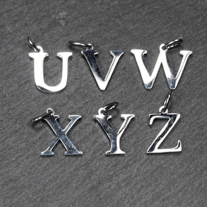 Sechs silberfarbene, hochglänzende Buchstaben-Anhänger (U V W X Y Z) in serifenförmiger Typografie mit kleiner Öse auf dunklem, strukturiertem Untergrund.