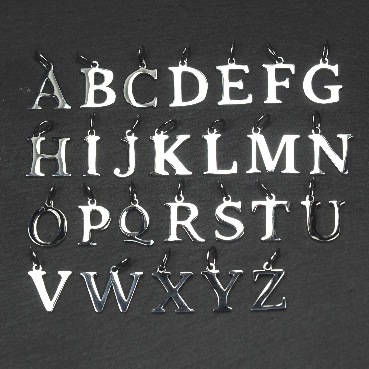 Buchstaben-Anhänger A–Z aus poliertem silberfarbenem Metall in serifenförmiger Typografie, mit Öse und Ring auf dunklem Grund.